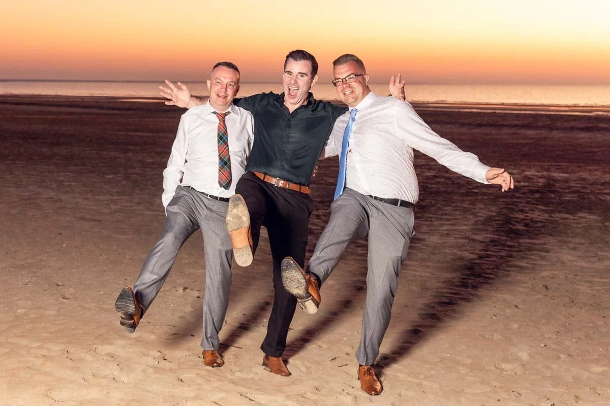 Three men in dress shirts and trousers joyfully dancing on a beach during sunset.