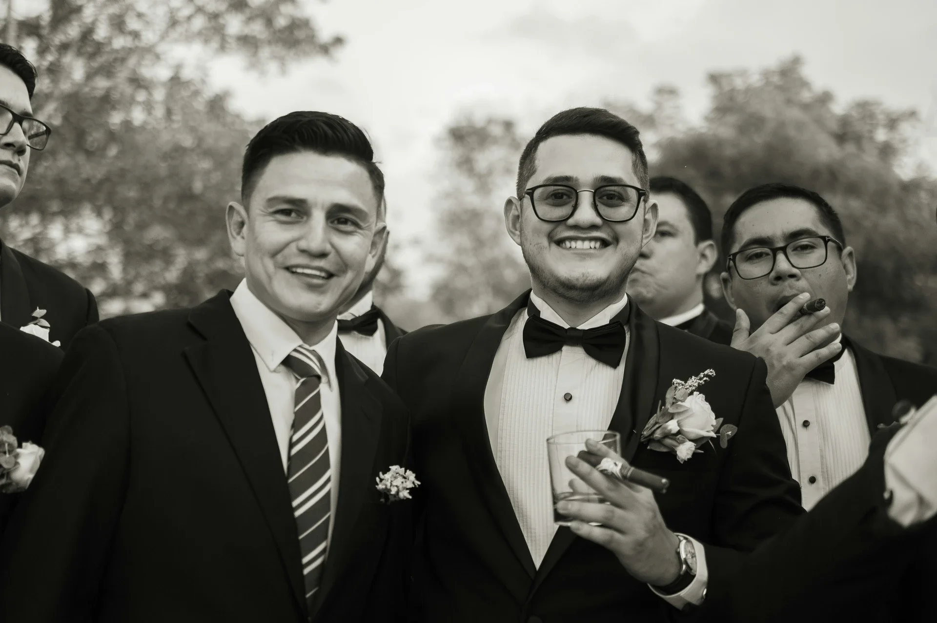 Group of men in tuxedos at formal event, smiling at camera, one holding a drink, outdoor setting with trees in background.