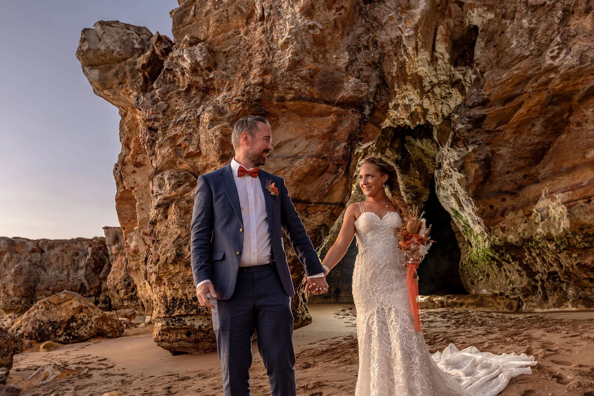 A newlywed couple holding hands on the beach, standing in front of a large rock formation at sunset. The groom is wearing a navy suit with an orange bow tie and boutonniere. The bride is in a white lace wedding dress holding a bouquet of dried flowers, and they are smiling at each other.