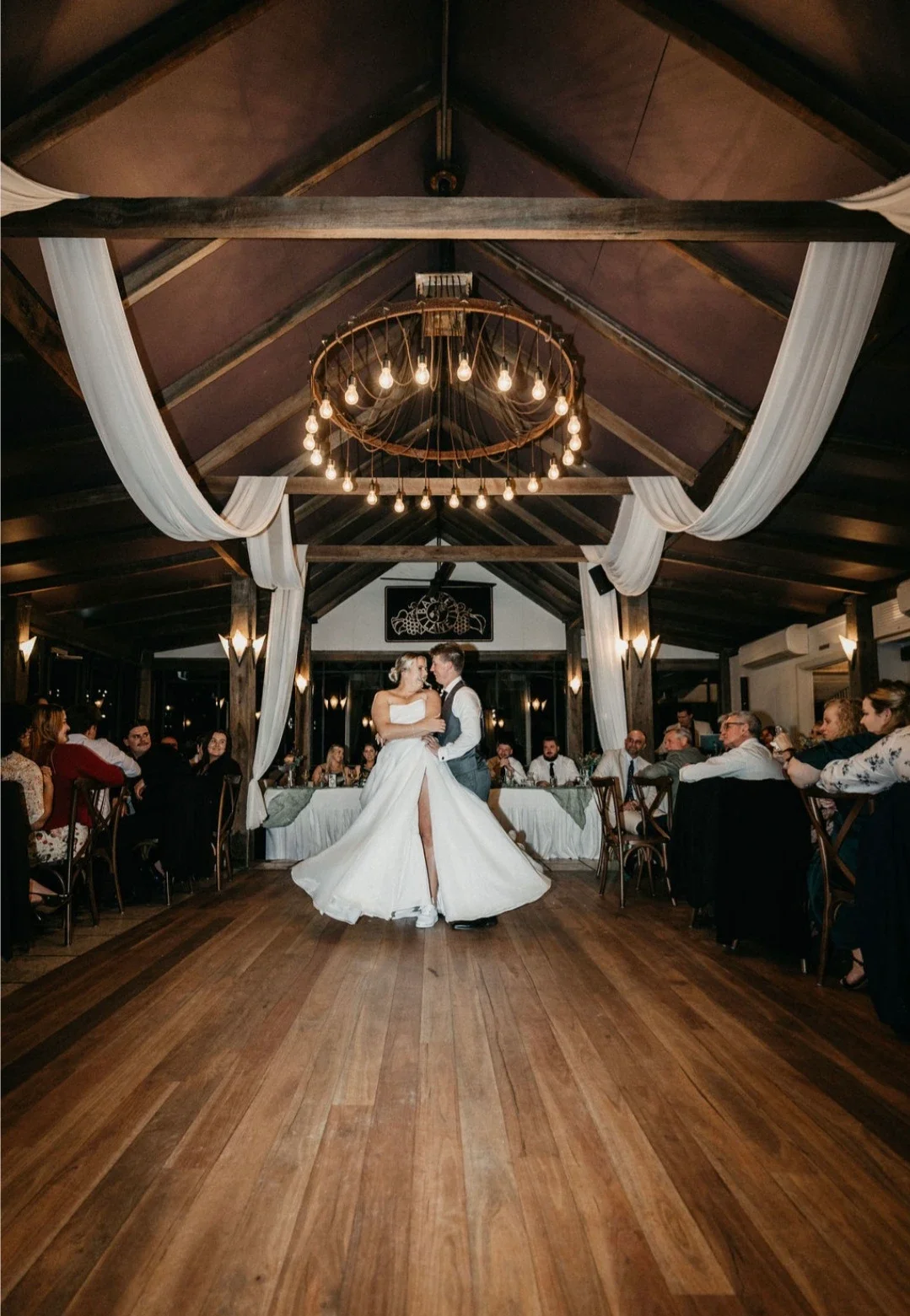 A bride and groom dancing in the center of a decorated reception hall with wooden floors and a high ceiling with draped white fabric and a circular chandelier with hanging light bulbs. Guests are seated around tables along the sides of the room.