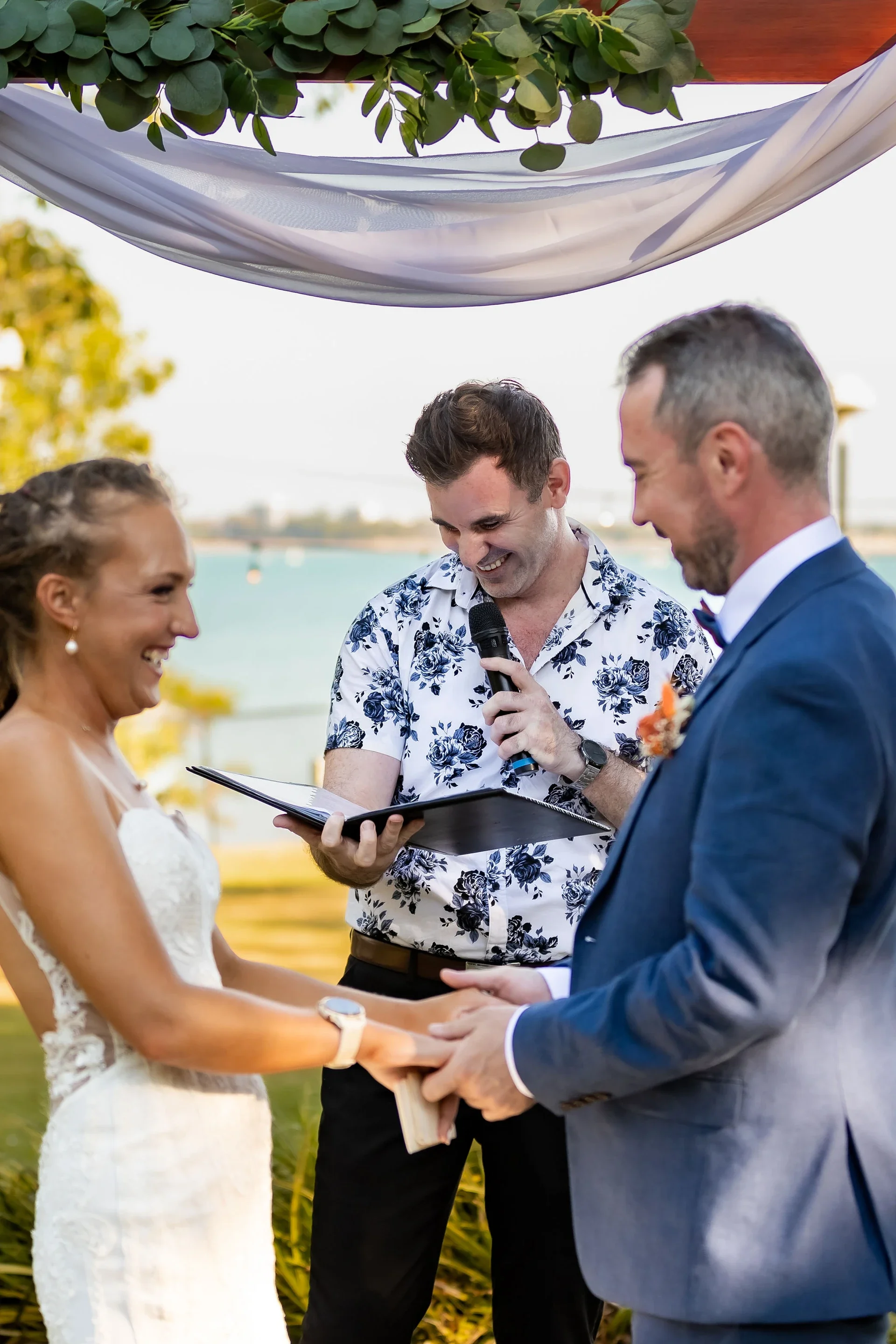 A wedding ceremony outdoors with a couple exchanging vows, an officiant reading from a book, against a scenic backdrop of water and trees.