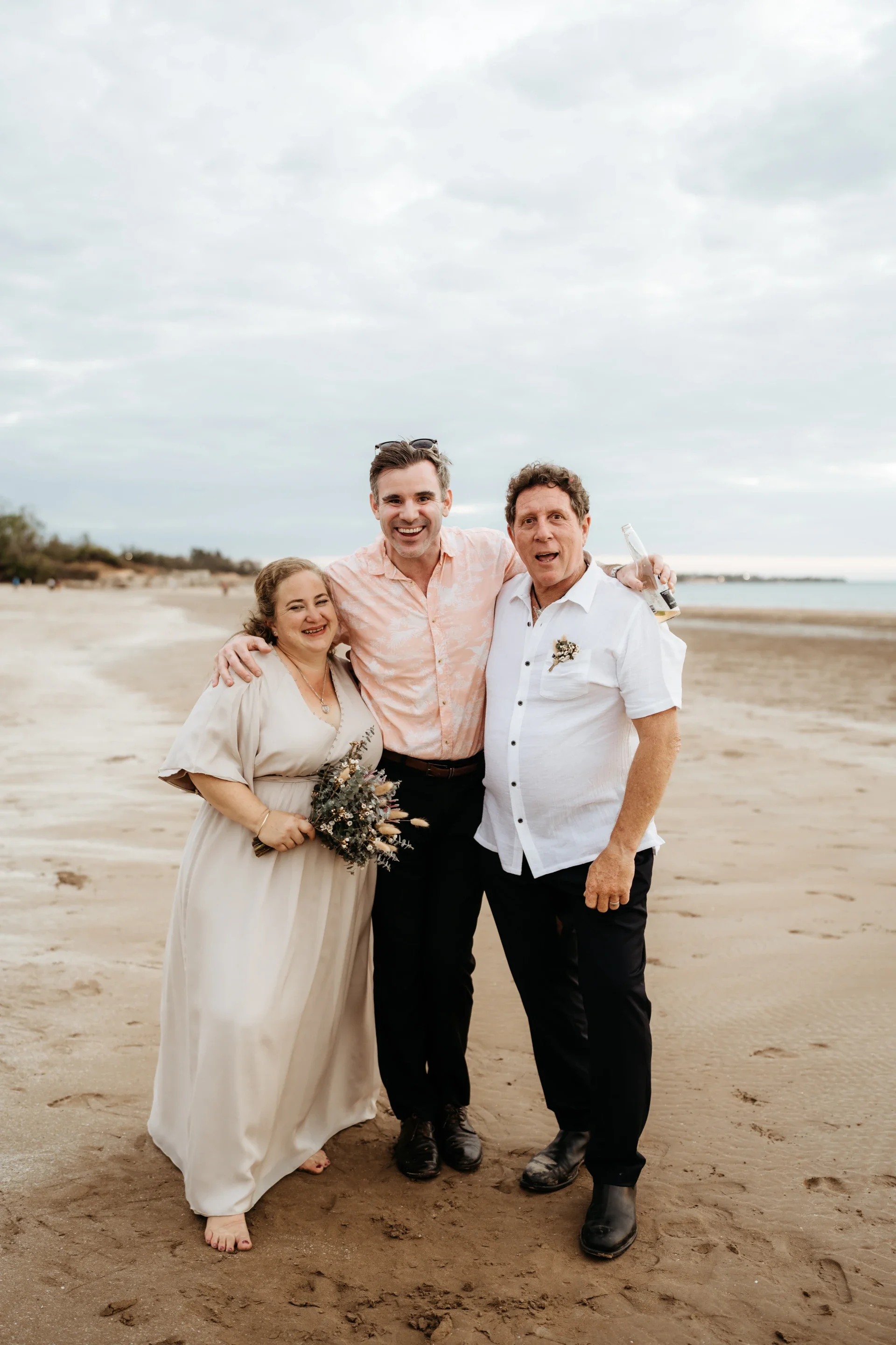 Three people standing on a sandy beach, smiling at the camera, with cloudy sky overhead. The woman on the left is holding a small bouquet, wearing a cream-colored dress. The man in the middle is wearing a light pink shirt, and the man on the right is