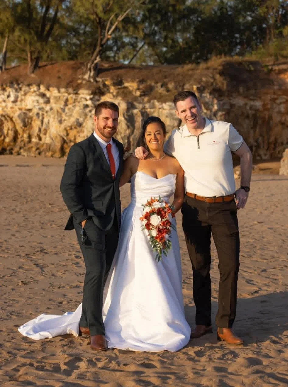 A bride in a white wedding dress holding a floral bouquet, standing on a sandy beach with two smiling men, one in a black suit and red tie, the other in a white polo with dark khaki pants, during sunset with rocky cliffs and trees in the background.