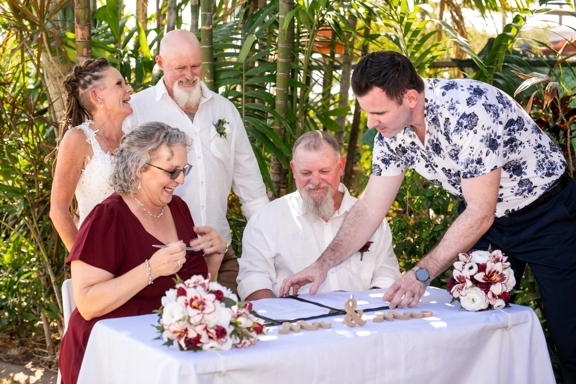 A group of six people celebrating a wedding outdoors, sitting and standing around a table with wedding documents and floral arrangements, with lush greenery in the background.