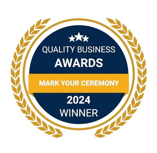 A circular award badge with a gold laurel wreath border, blue center, and a yellow banner reading 'Mark Your Ceremony.' Text on the badge reads 'Quality Business Awards 2024 Winner.'
