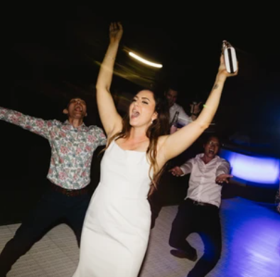 Group of people dancing at a party with a woman in a white dress holding a drink and raising her arm