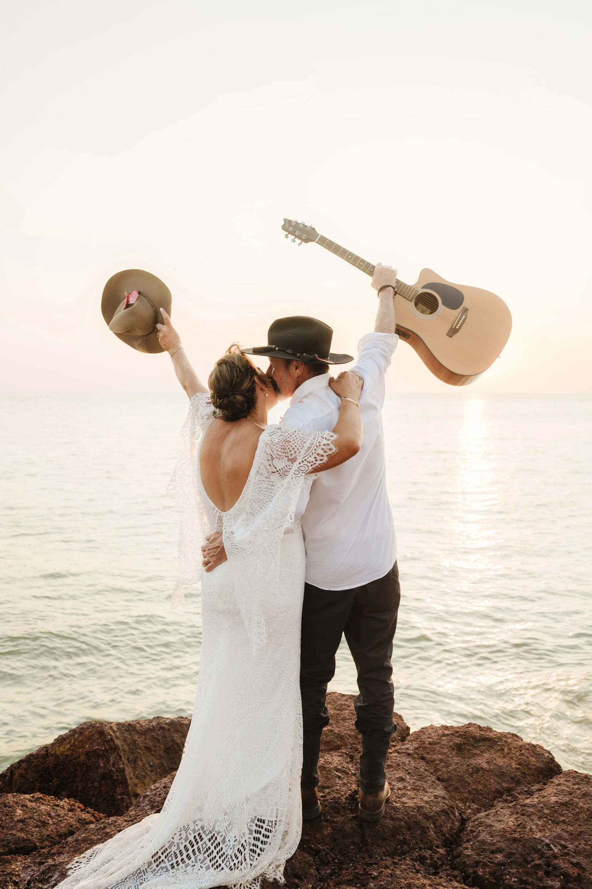 Couple embracing on rocks by the water at sunset, with the woman in a white lace dress and the man in a white shirt and black hat. They are holding a hat and a guitar above their heads, sharing a kiss.