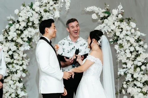 Bride and groom holding hands during wedding ceremony with officiant, surrounded by white floral arrangements.