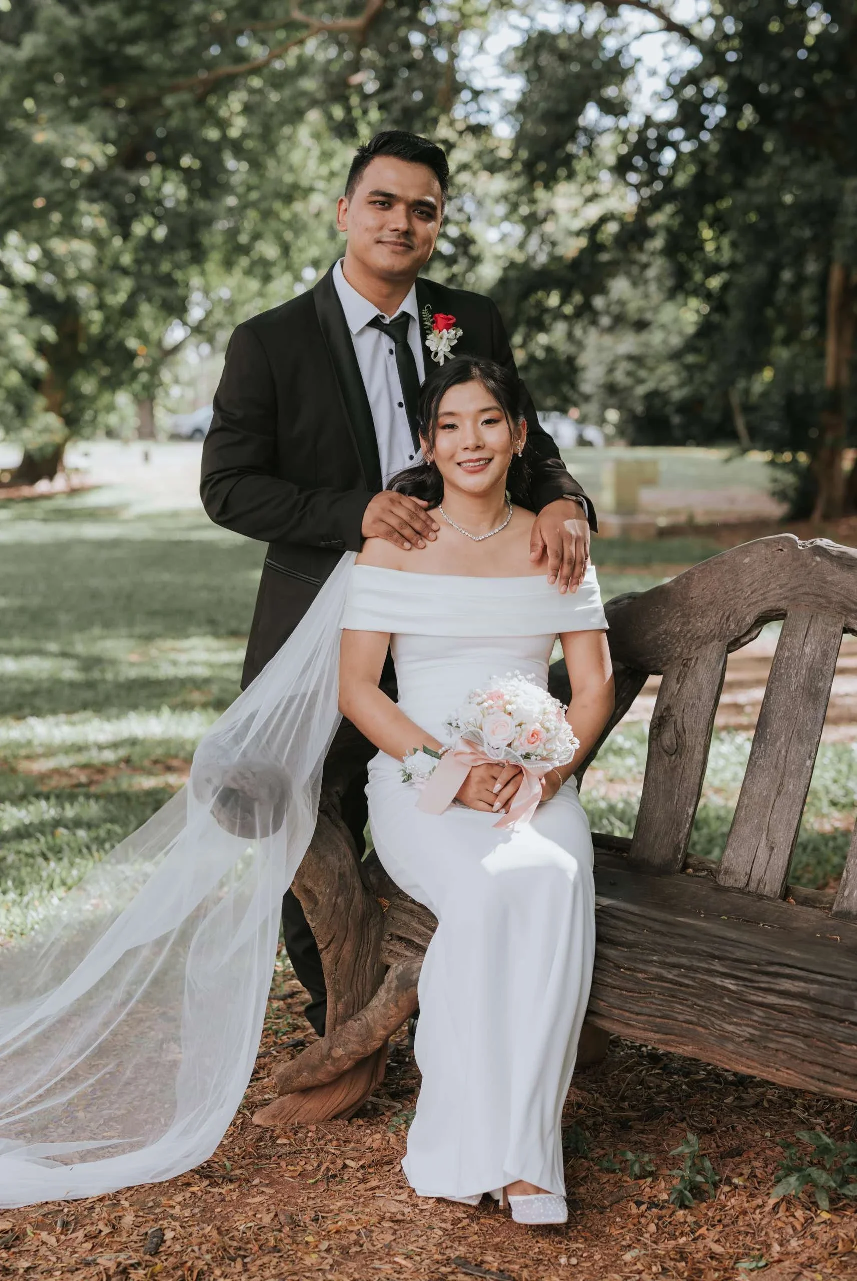 A newlywed couple outdoors, the bride in a white off-shoulder gown with a veil and holding a bouquet of pink and white flowers, sitting on a wooden bench, the groom in a black suit with a white shirt and black tie standing behind her with his hand on her shoulder, in a park surrounded by trees and greenery.