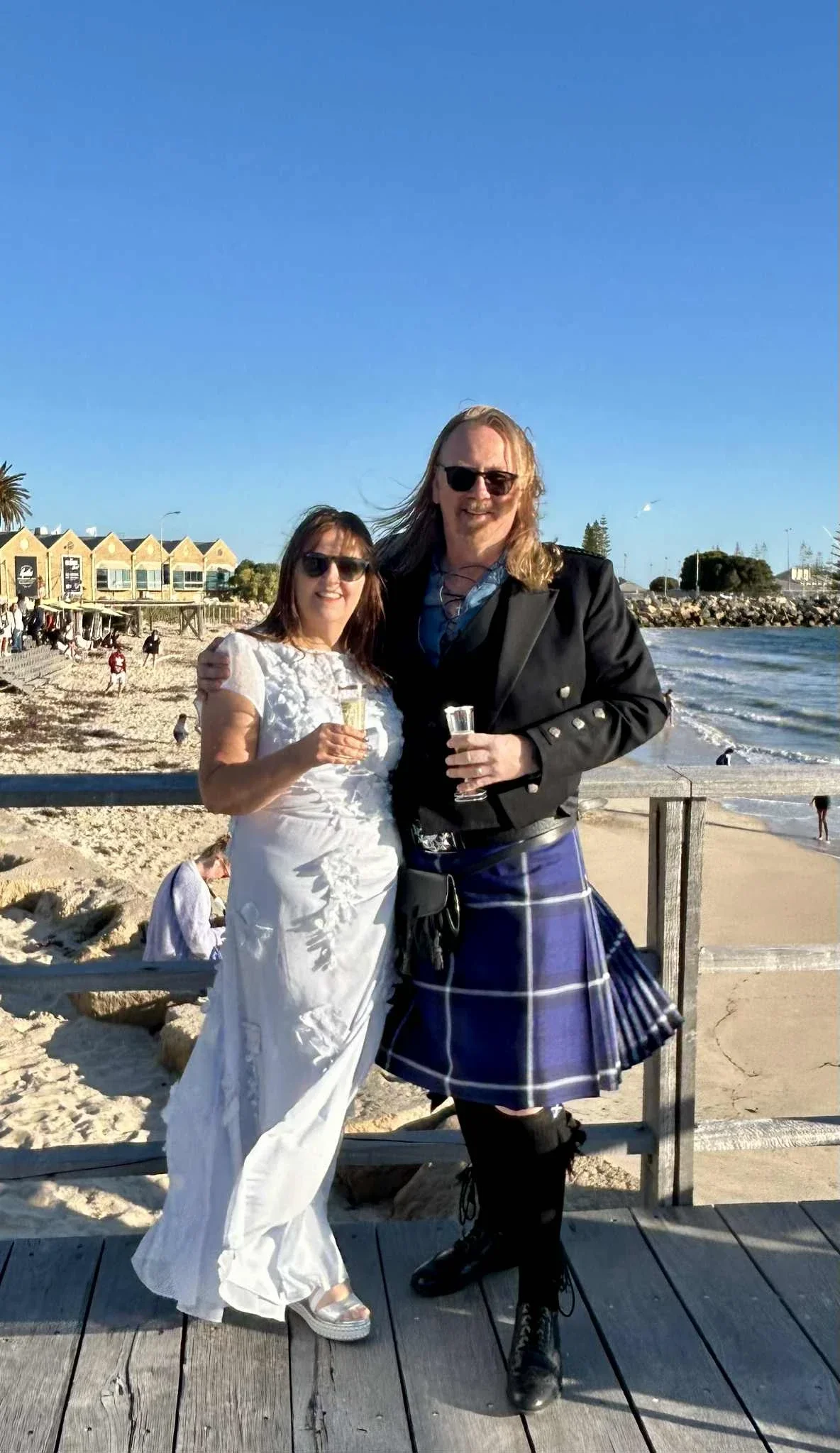 Two people standing on a wooden dock by the beach, smiling, holding champagne glasses, with the ocean and a clear blue sky in the background. One person is wearing a kilt and black boots, and the other person is wearing a white dress and silver shoes