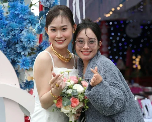 Two women smiling, one in a white dress holding a bouquet of pink and white flowers, and the other in a gray jacket making a finger heart gesture, standing in front of blue and purple festive decorations.