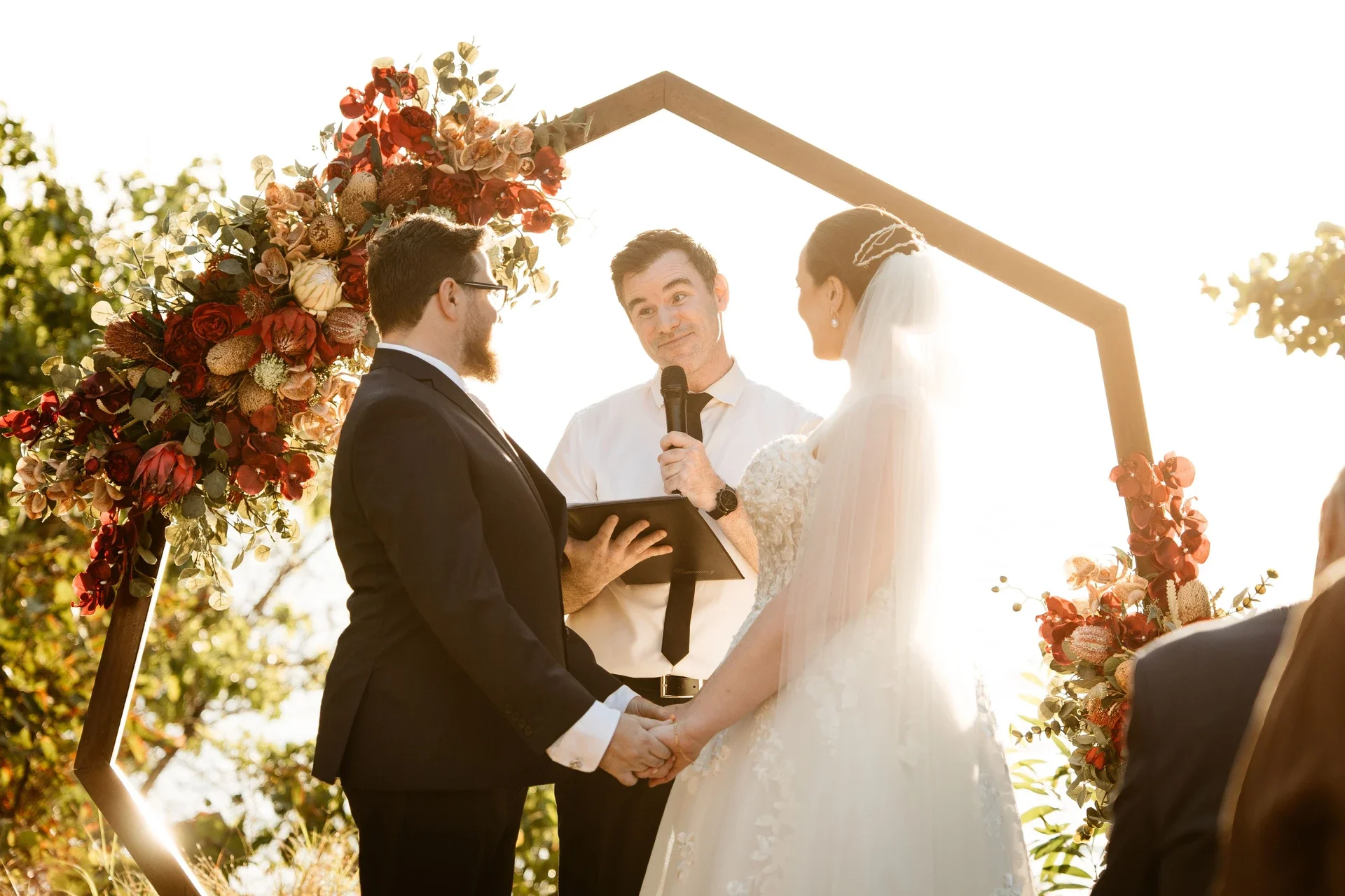 A wedding ceremony outdoors with a couple holding hands and an officiant in the background, surrounded by floral decorations and sunlight.