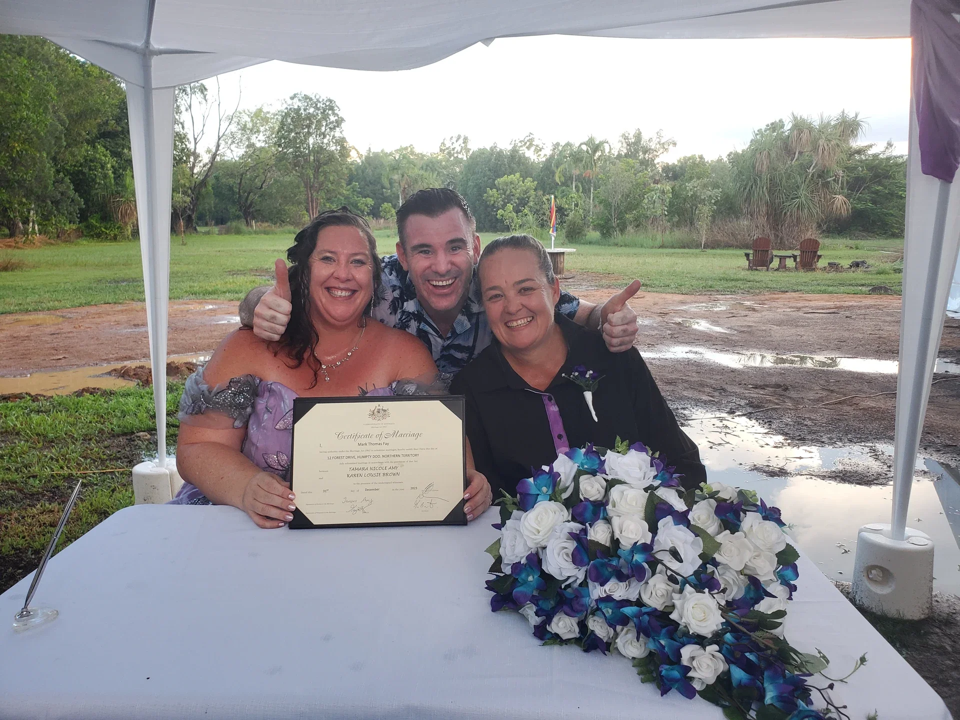 Three people smiling and celebrating a marriage outdoors under a white tent, with a certificate of marriage and a bouquet of white and purple flowers on the table in front of them.
