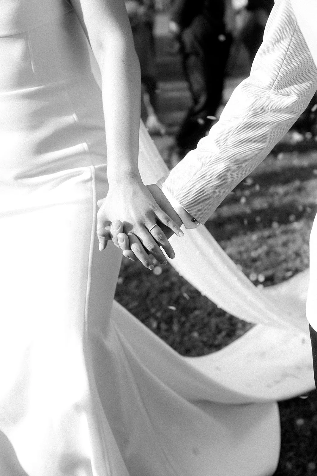 Close-up of a bride and groom holding hands during a wedding, with the bride wearing a satin wedding gown and the groom in a textured suit jacket, outdoors in black and white.