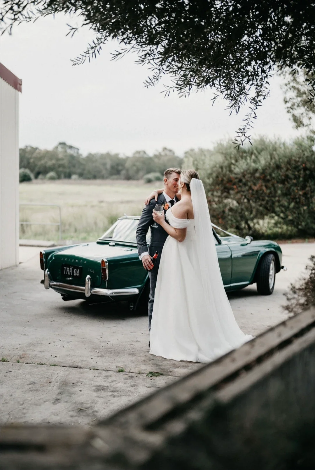 A bride and groom in wedding attire sharing a kiss outdoors near a vintage green car, with nature and trees in the background.
