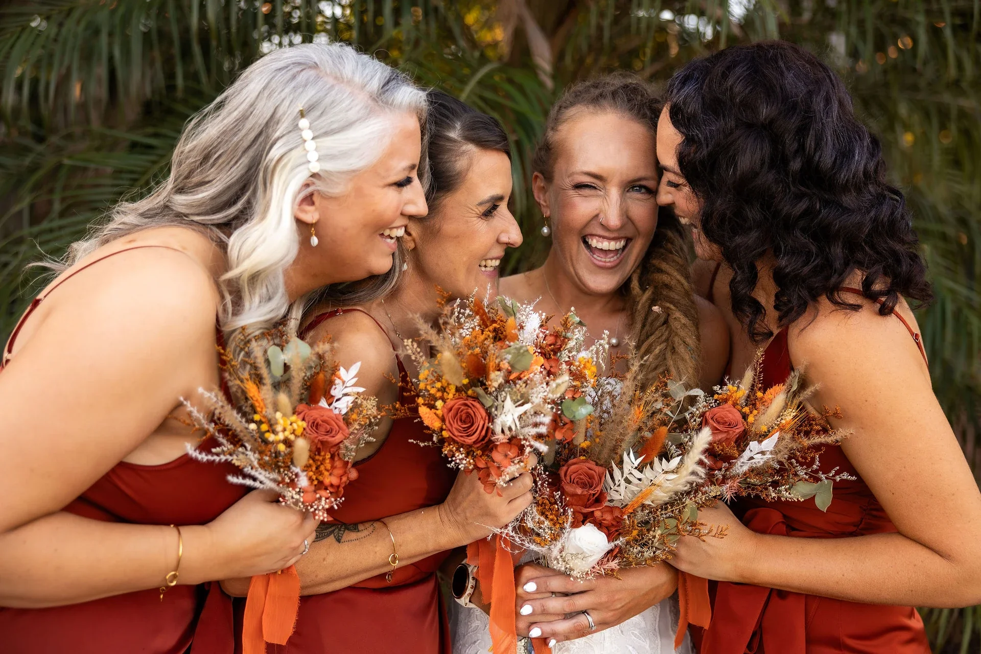 Four women celebrating a wedding, dressed in matching rust-colored dresses, holding bouquets of orange, white, and earthy-toned flowers, smiling and laughing closely together outdoors.