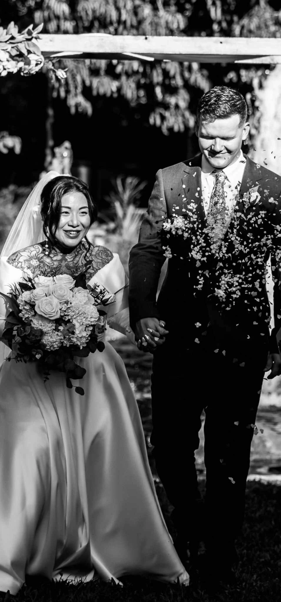 A black and white photo of a bride and groom walking hand in hand outdoors, with flowers and greenery around, celebrating their wedding.