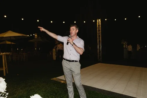 A young man in a white shirt and gray pants speaking into a microphone at an outdoor evening event, with string lights and other people in the background.