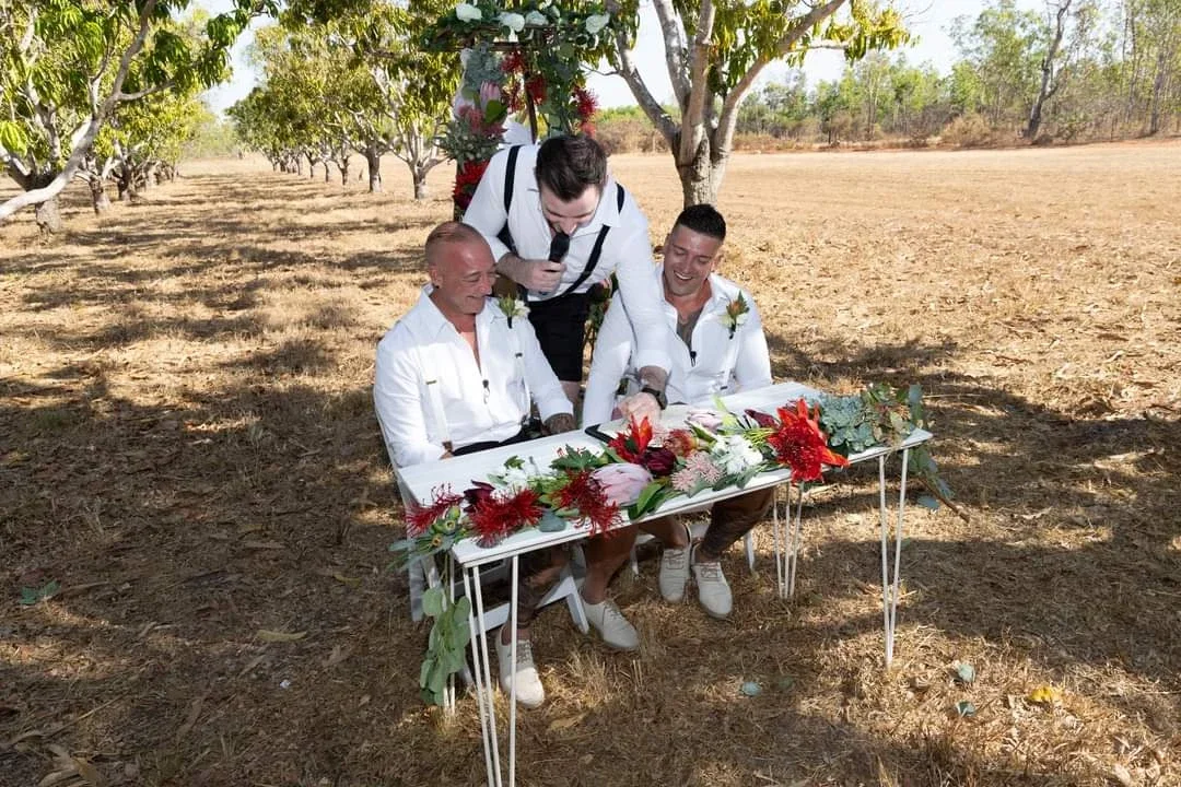 Three men participating in an outdoor wedding ceremony, two sitting at a table decorated with flowers and one standing, speaking into a microphone. The scene is set under a tree in a rural area.