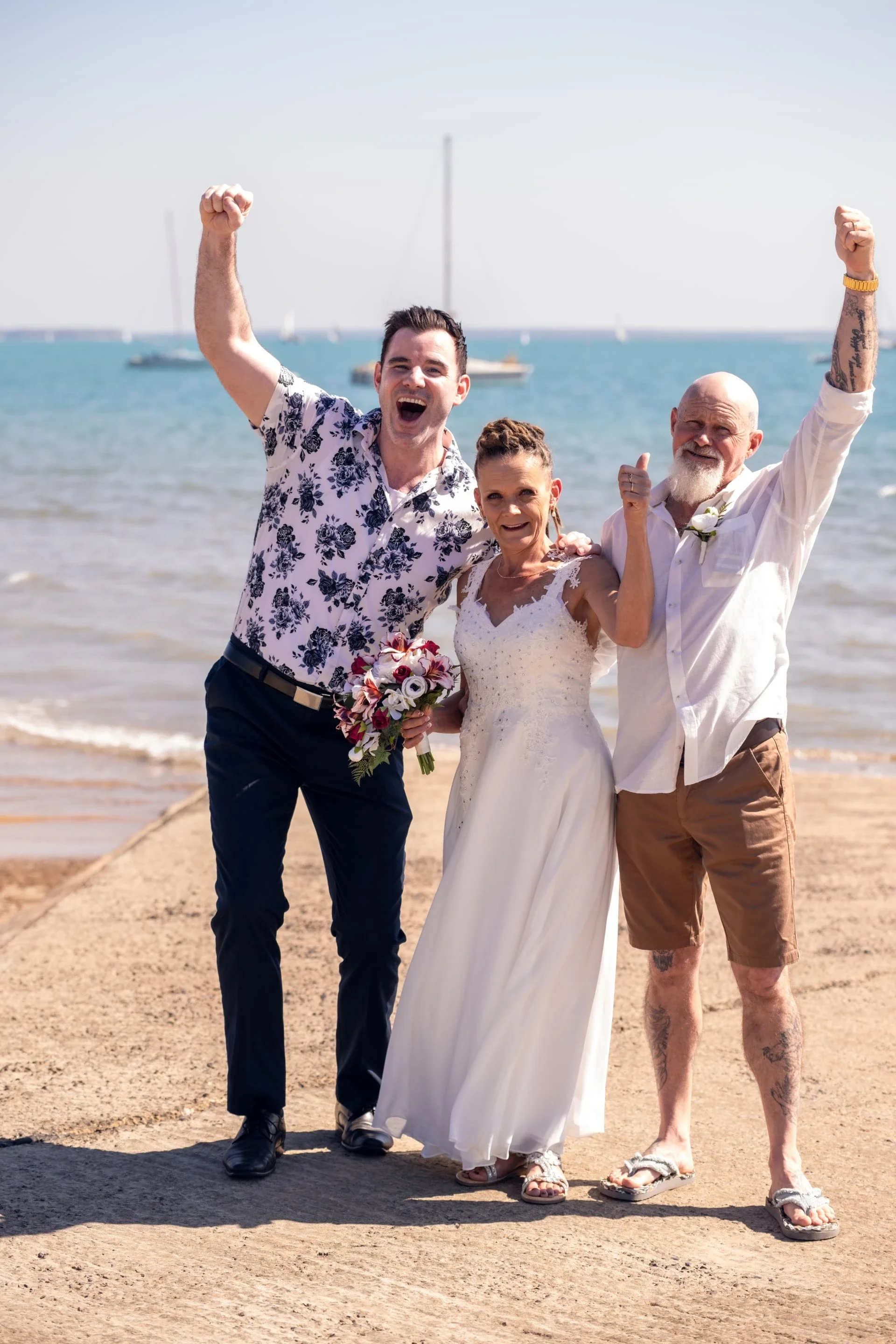Three people, including a bride in a white wedding dress, celebrating on a beach with arms raised, with sailboats in the background.