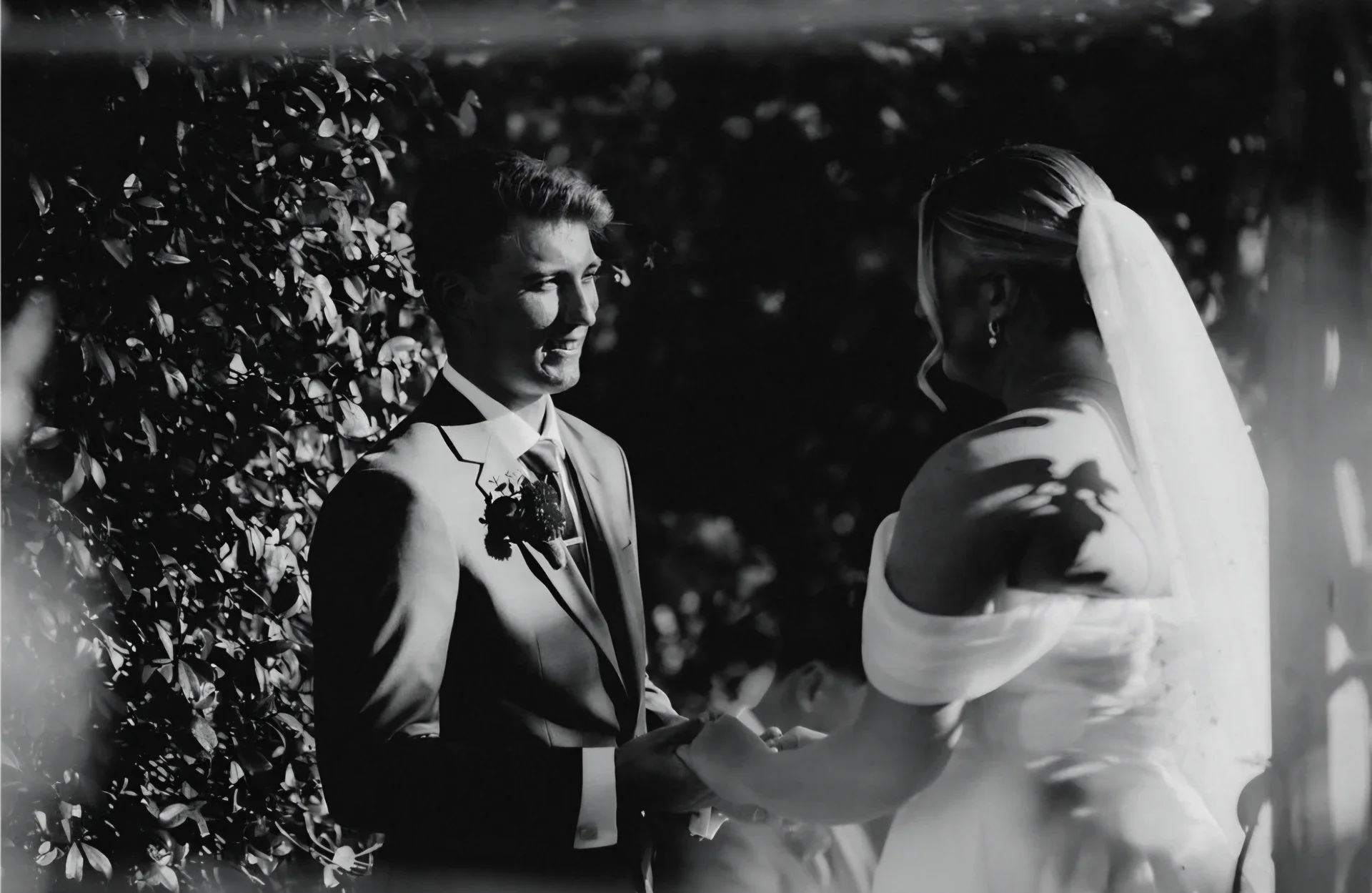 A black and white photograph of a bride and groom holding hands outdoors during their wedding ceremony, with the groom smiling at the bride, and shadows of leaves cast on the bride's face and shoulder.