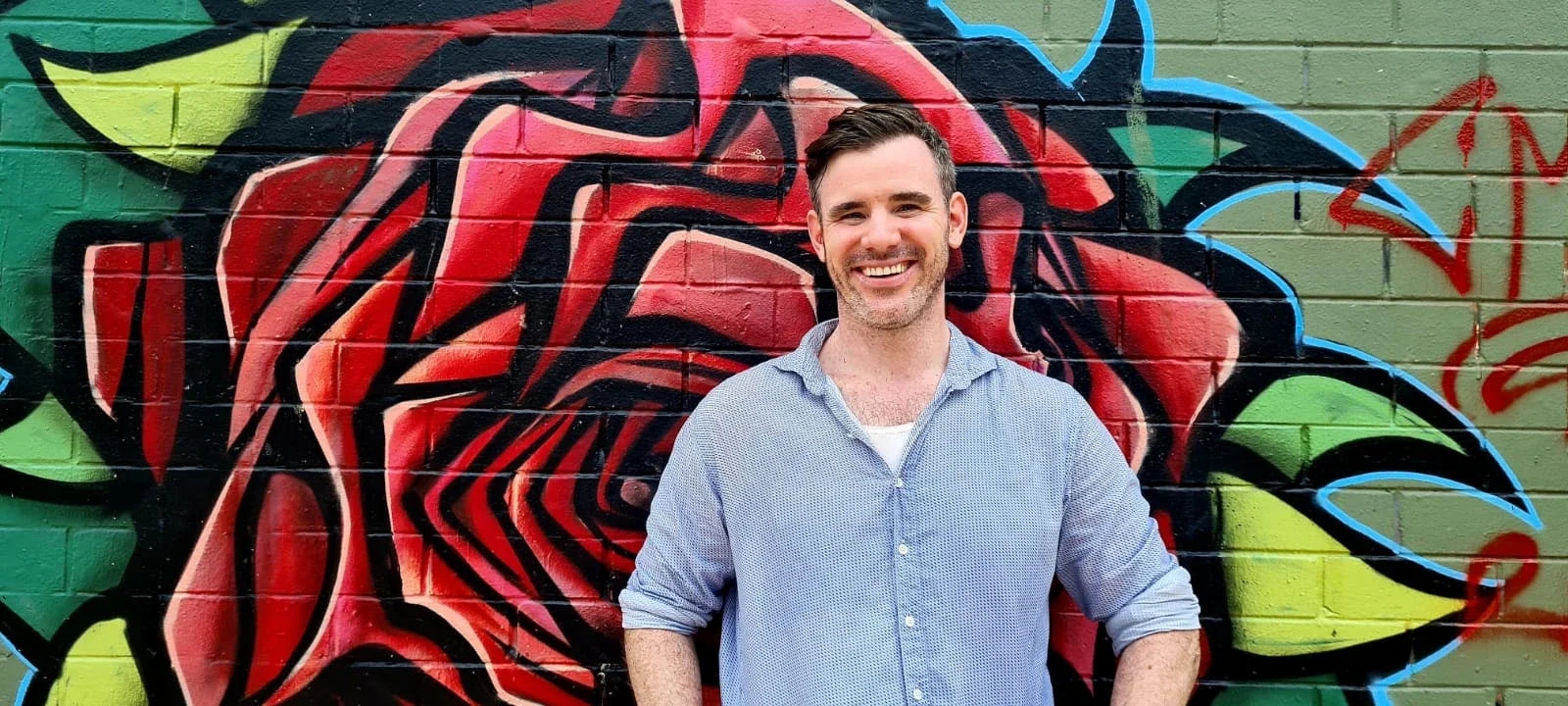 A man with short dark hair and a beard, smiling, wearing a light blue button-up shirt, standing in front of colorful graffiti art depicting a rose with green, red, black, yellow, and blue accents.
