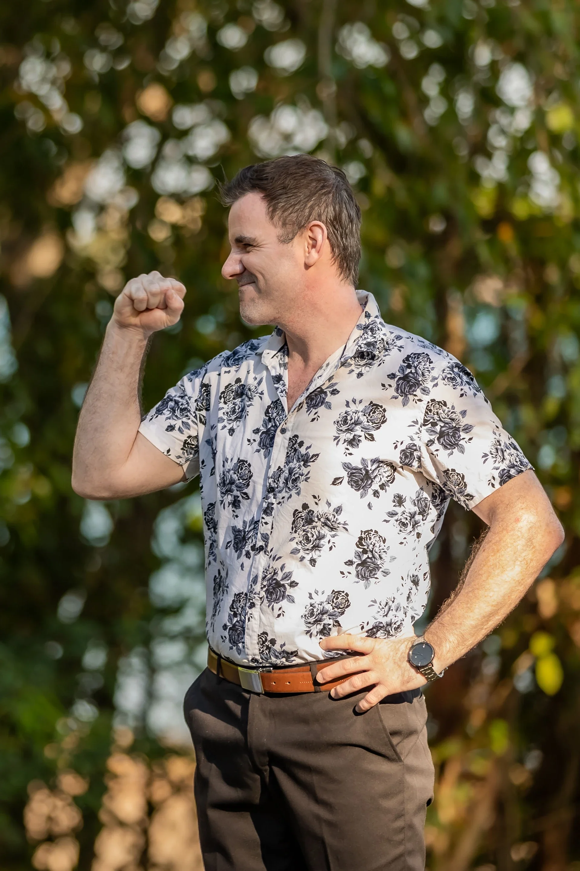 A man in a floral shirt making a victorious gesture with a clenched fist, smiling, outdoors with trees in the background.