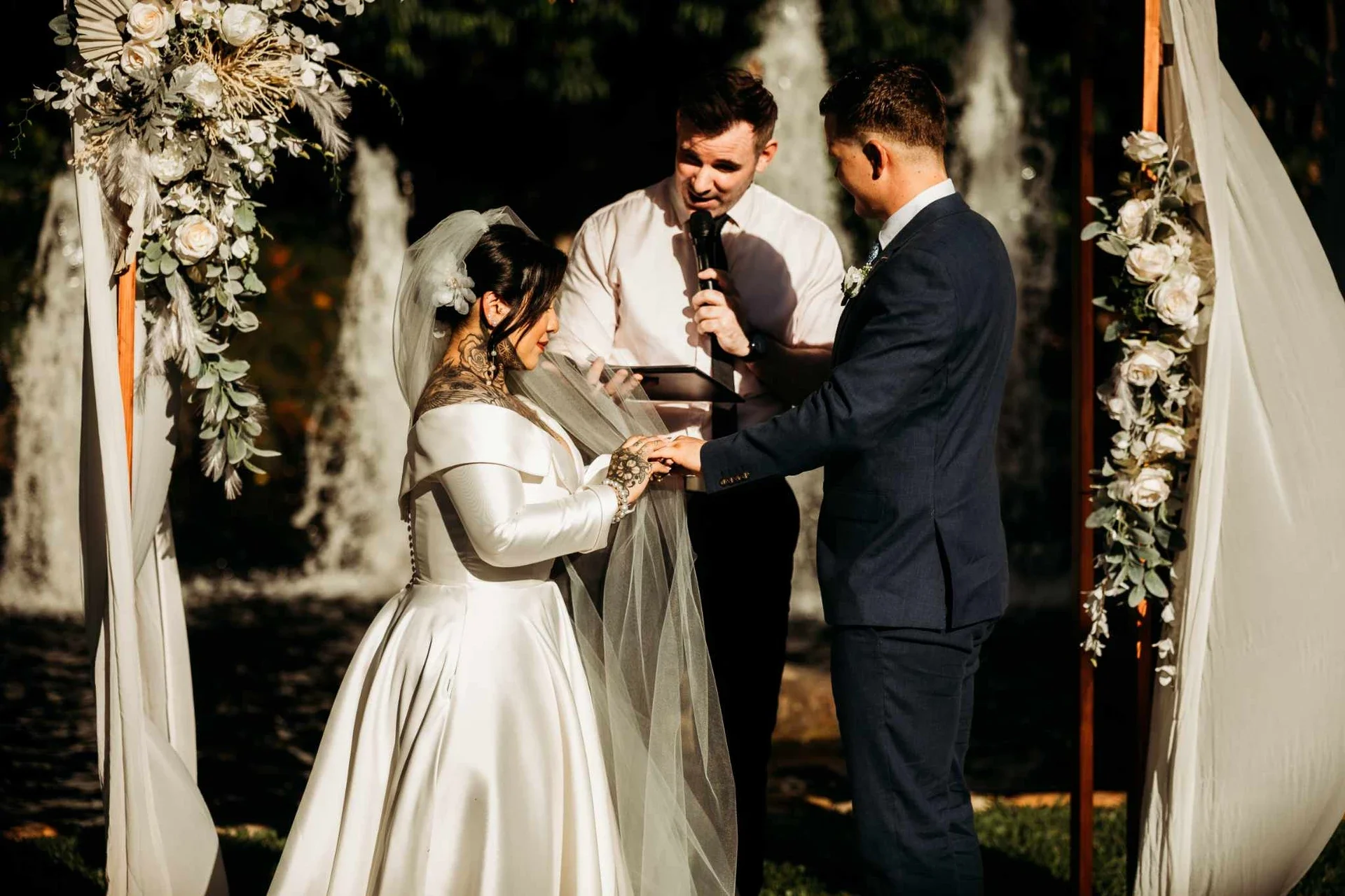 A wedding ceremony taking place outdoors with a bride and groom exchanging rings under a floral arch, officiant reading from a tablet, during daylight.