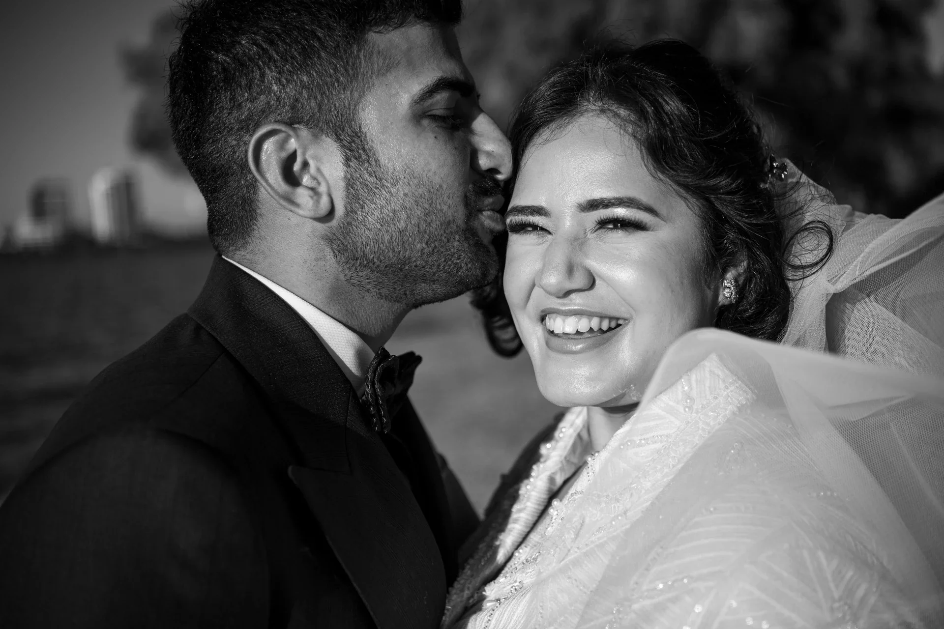 A black and white photograph of a man and woman on their wedding day. The man, in a tuxedo, is gently kissing the woman on her forehead. The woman, smiling and showing her teeth, is dressed in a wedding gown with lace details and tiara earrings. The 