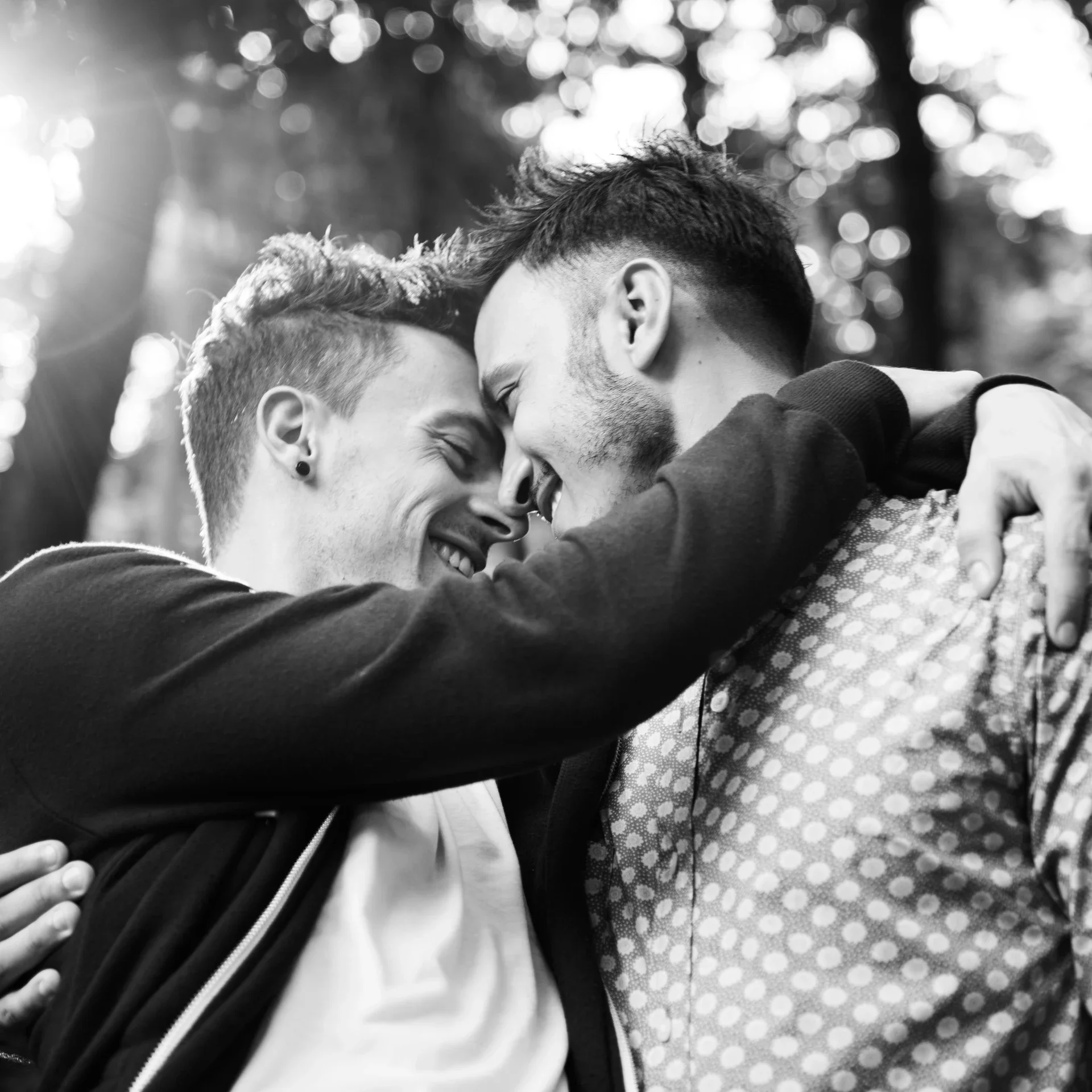 Two young men are smiling and hugging each other closely outdoors, with sunlight filtering through trees in the background.