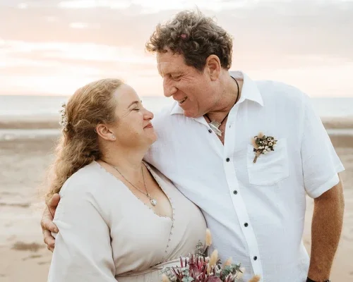A couple smiling at each other on a beach at sunset, holding a bouquet of flowers.