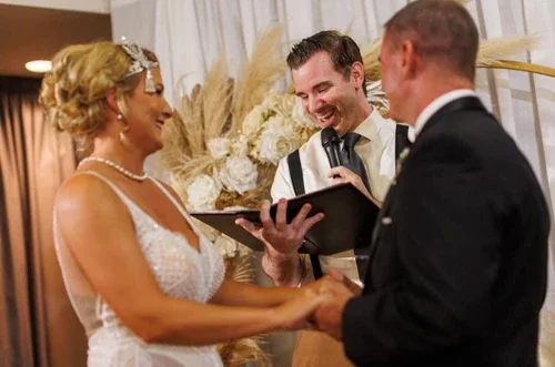 A bride and groom holding hands during their wedding ceremony as an officiant reads from a book. The bride is dressed in a white gown and the groom in a tuxedo. They are smiling at each other in a decorated indoor setting.