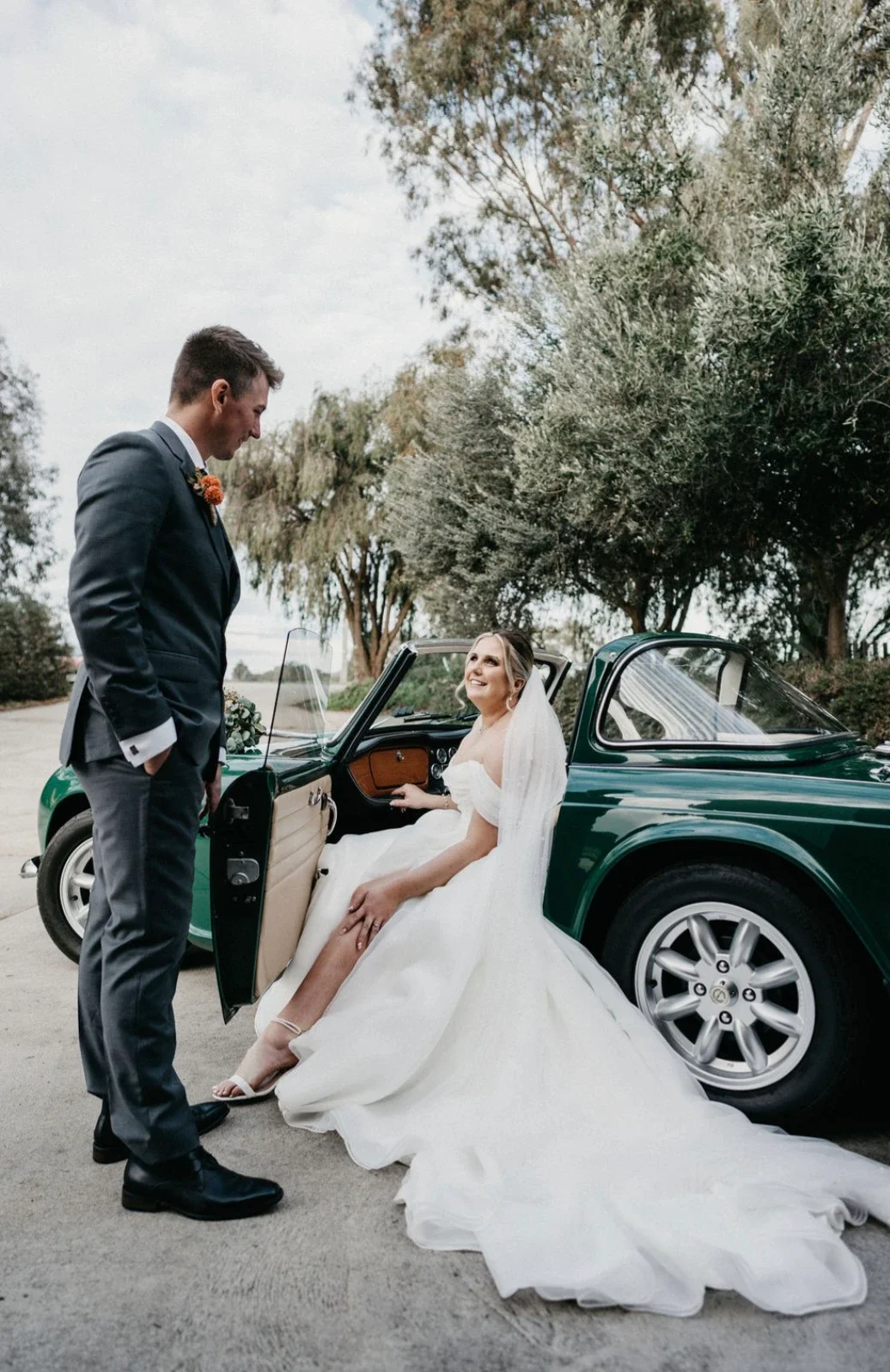 A bride sitting in a vintage green car with a white wedding dress, smiling at a groom in a suit, outside in a park with trees.