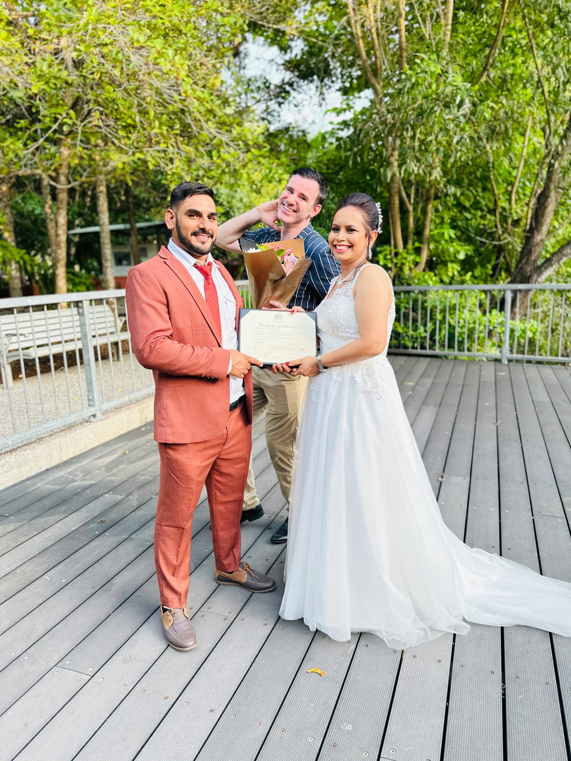 Wedding couple with a man in a stylish suit and a woman in a white wedding dress, holding a marriage certificate, smiling outdoors with a man delivering flowers in the background.