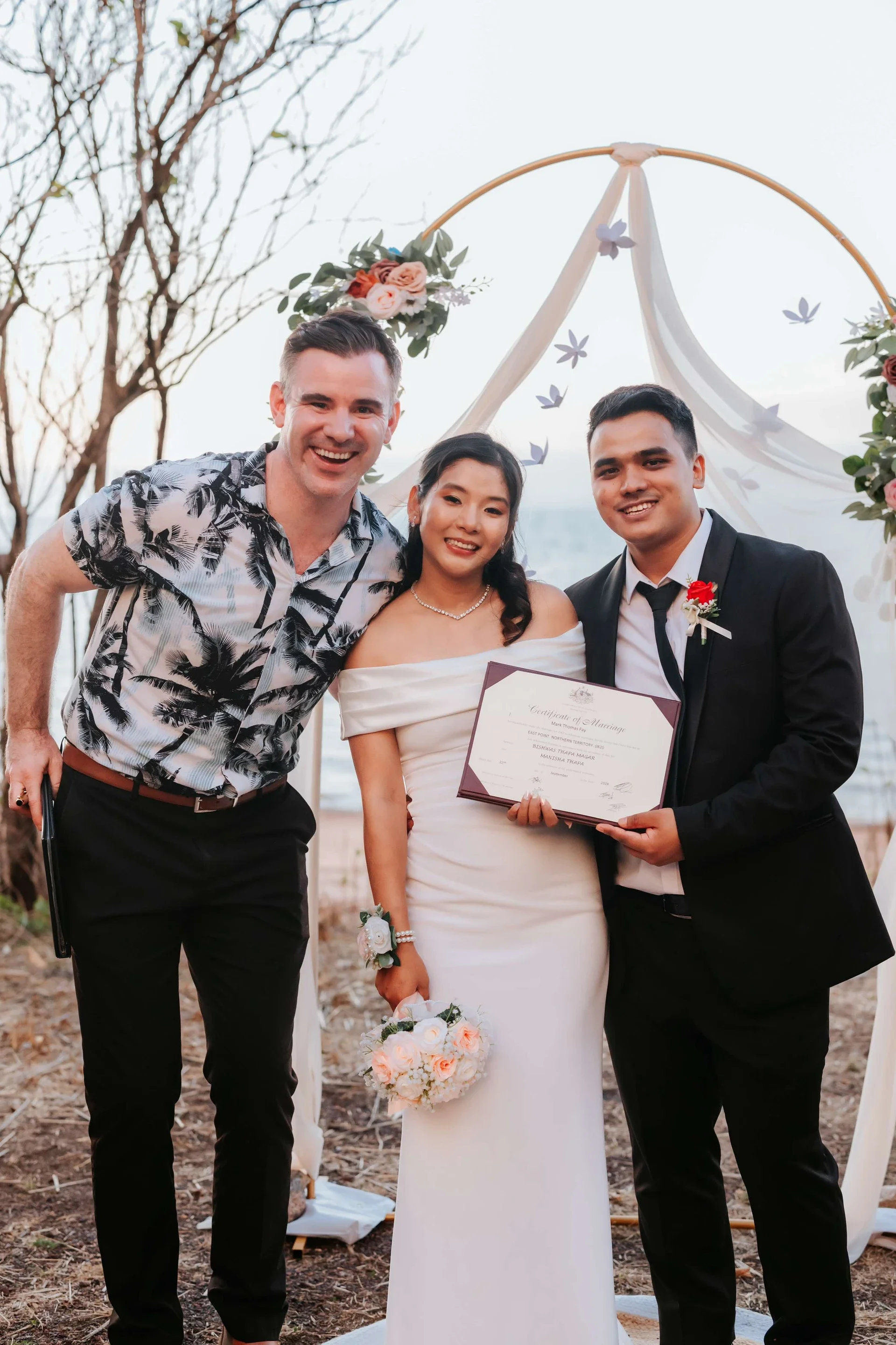 A wedding ceremony with two men and a woman standing under a decorative arch, with the man on the right holding a marriage certificate, in an outdoor setting during sunset.