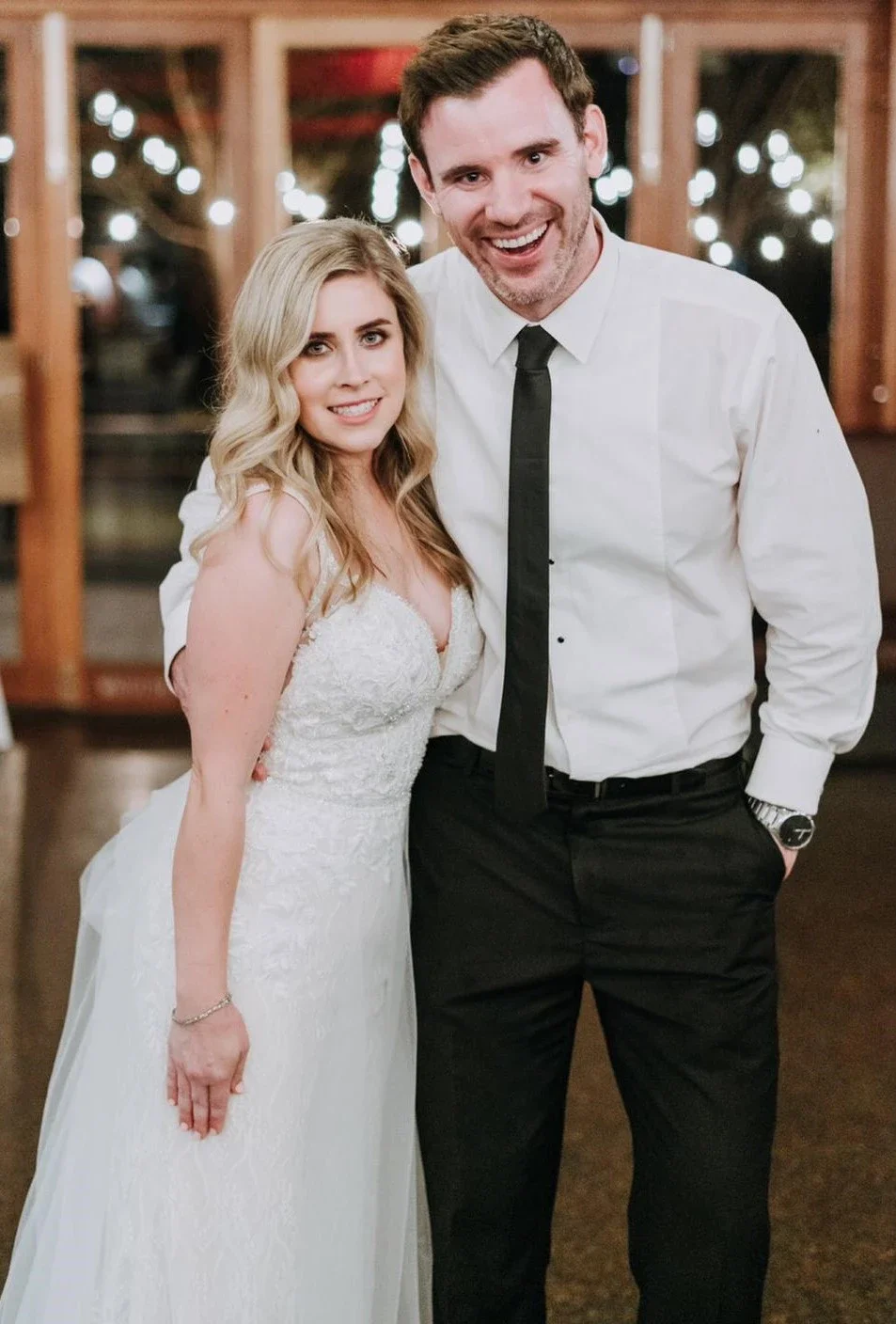A bride in a white wedding dress and a groom in a white shirt with a black tie standing together indoors at a wedding reception, smiling at the camera.