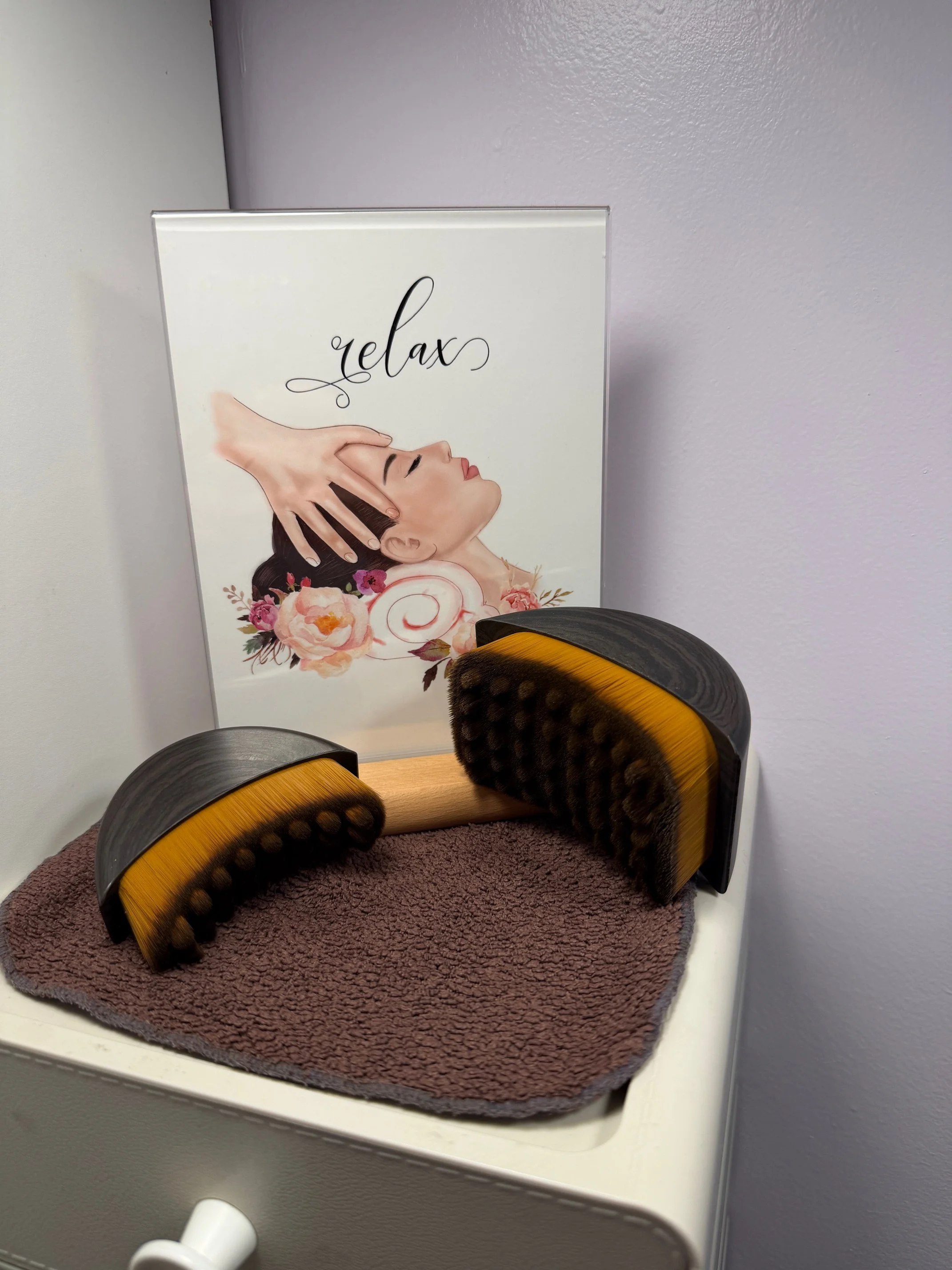 A massage roller with black and yellow bristles resting on a brown towel on top of a white surface, with a sign in the background showing a woman receiving a massage with the word 'relax' written above.