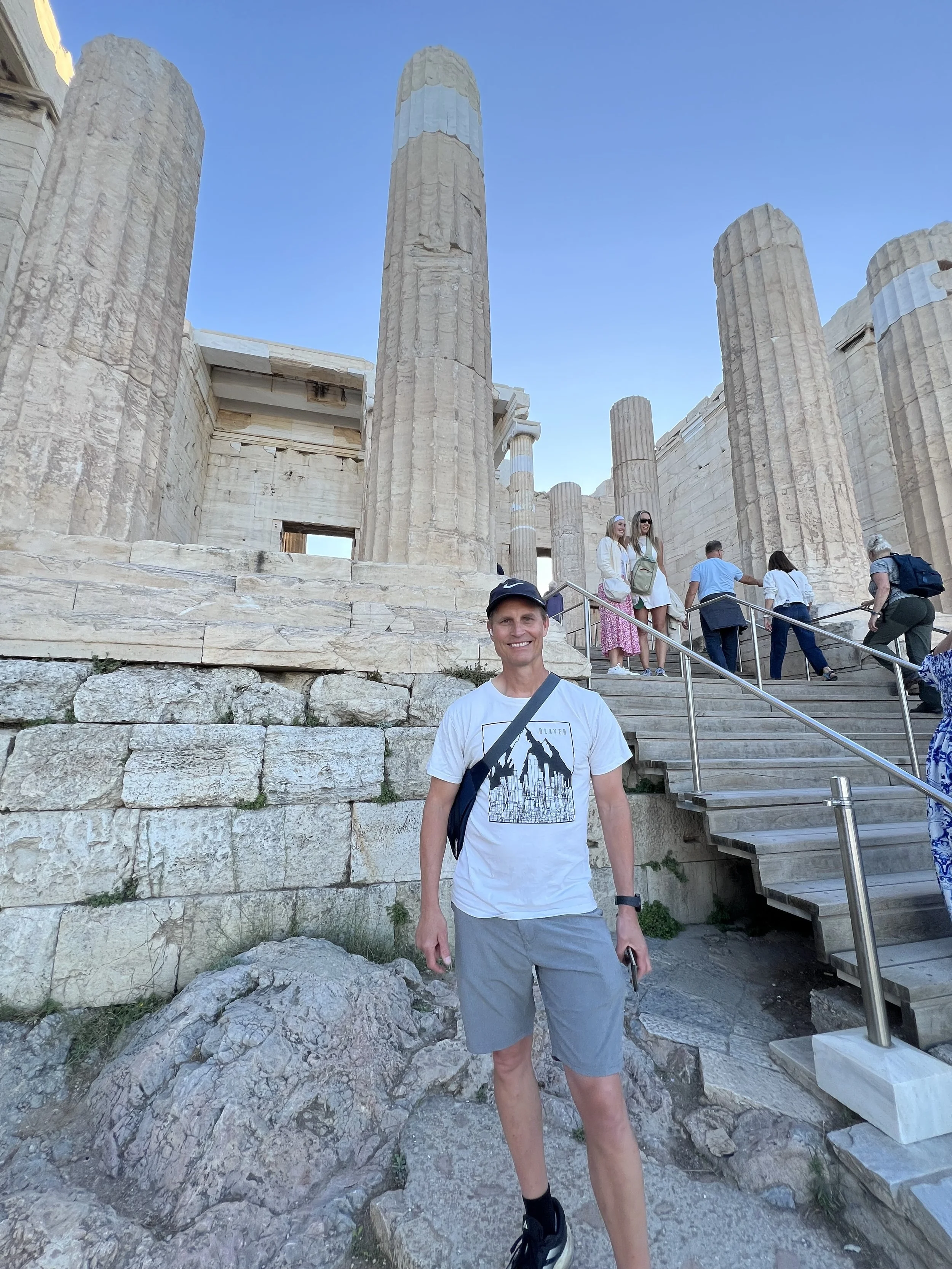 Man in white t-shirt and grey shorts smiling at the camera, standing in front of ancient Greek ruins with large stone columns and stairs, with other visitors ascending the stairs behind him.