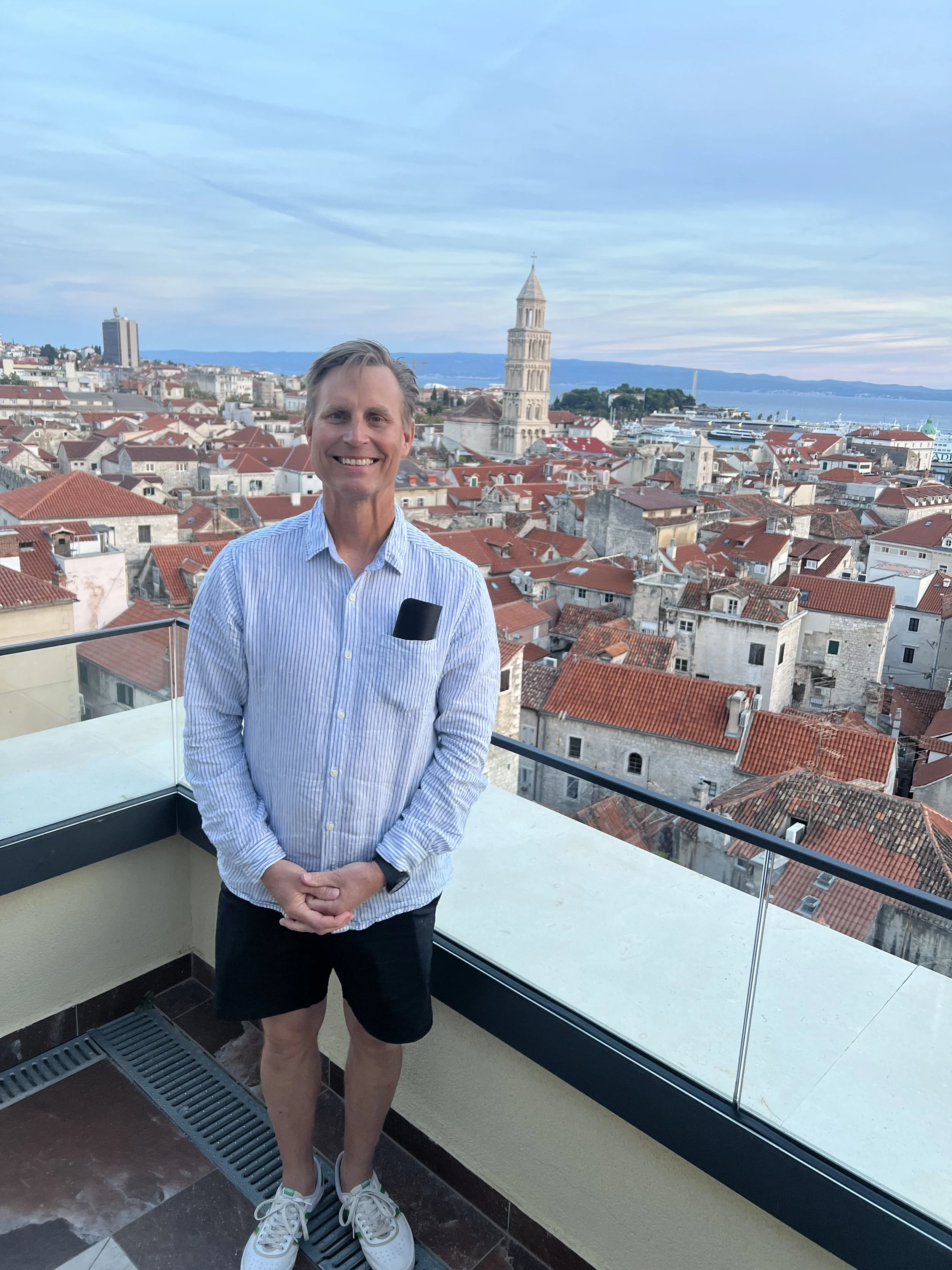 Man smiling outdoors on a rooftop with a cityscape featuring red-tiled roofs, a church tower, and water in the background.