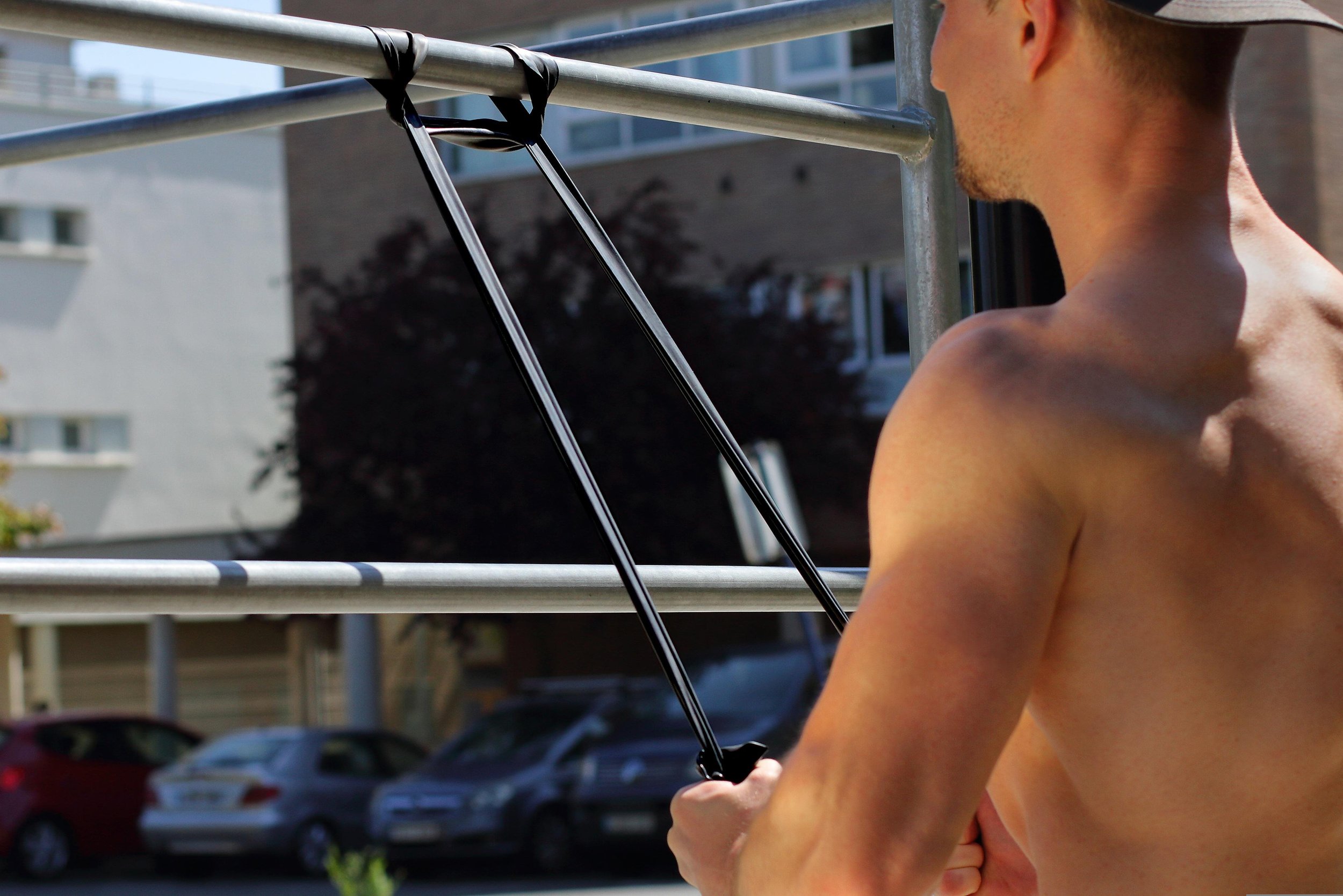 A man with a muscular build and no shirt is holding resistance bands attached to a metal structure outdoors doing shoulder strength exercises