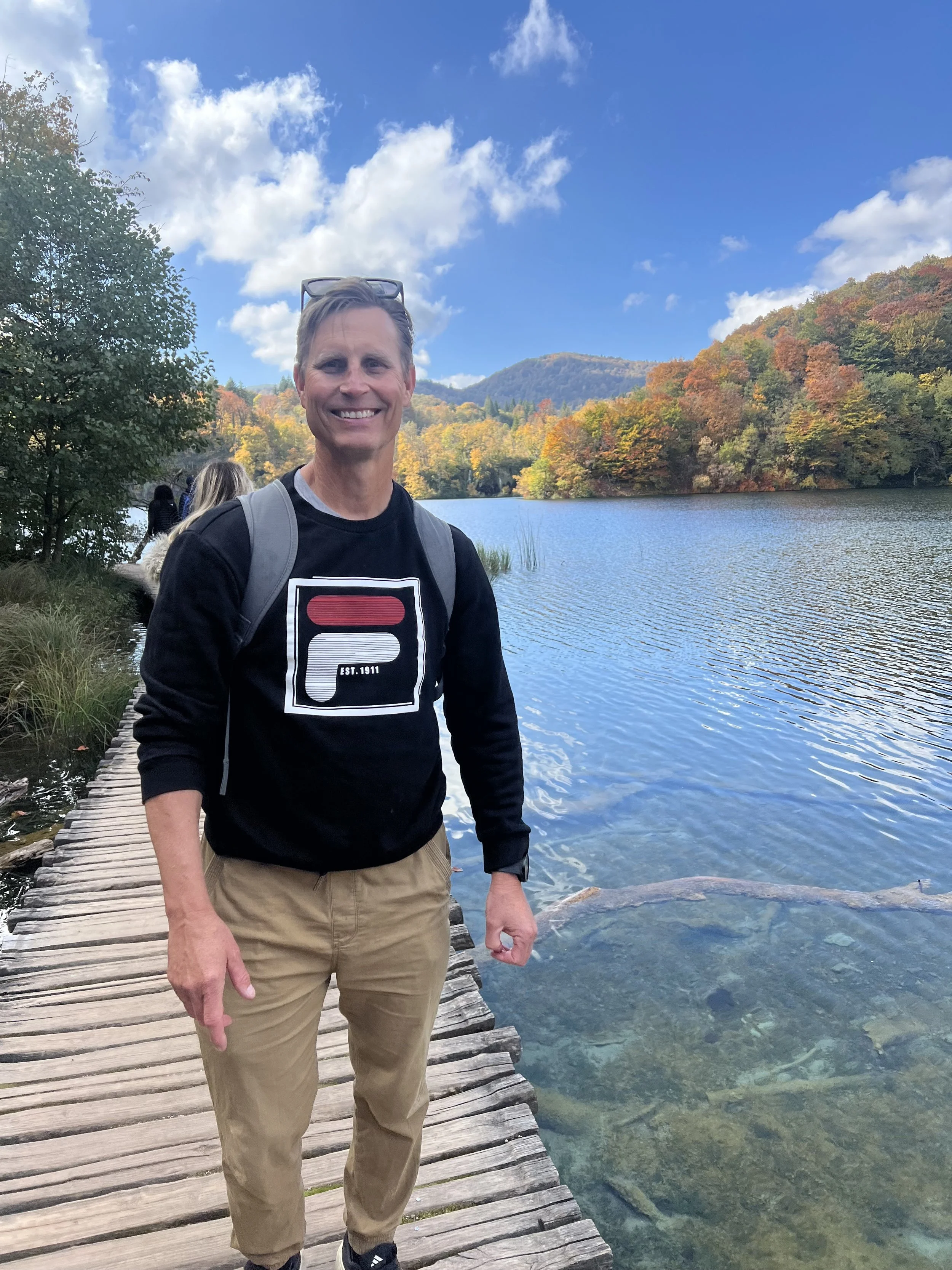 A smiling man in a black sweatshirt with a logo, tan pants, and a backpack standing on a wooden dock beside a lake surrounded by colorful autumn trees.