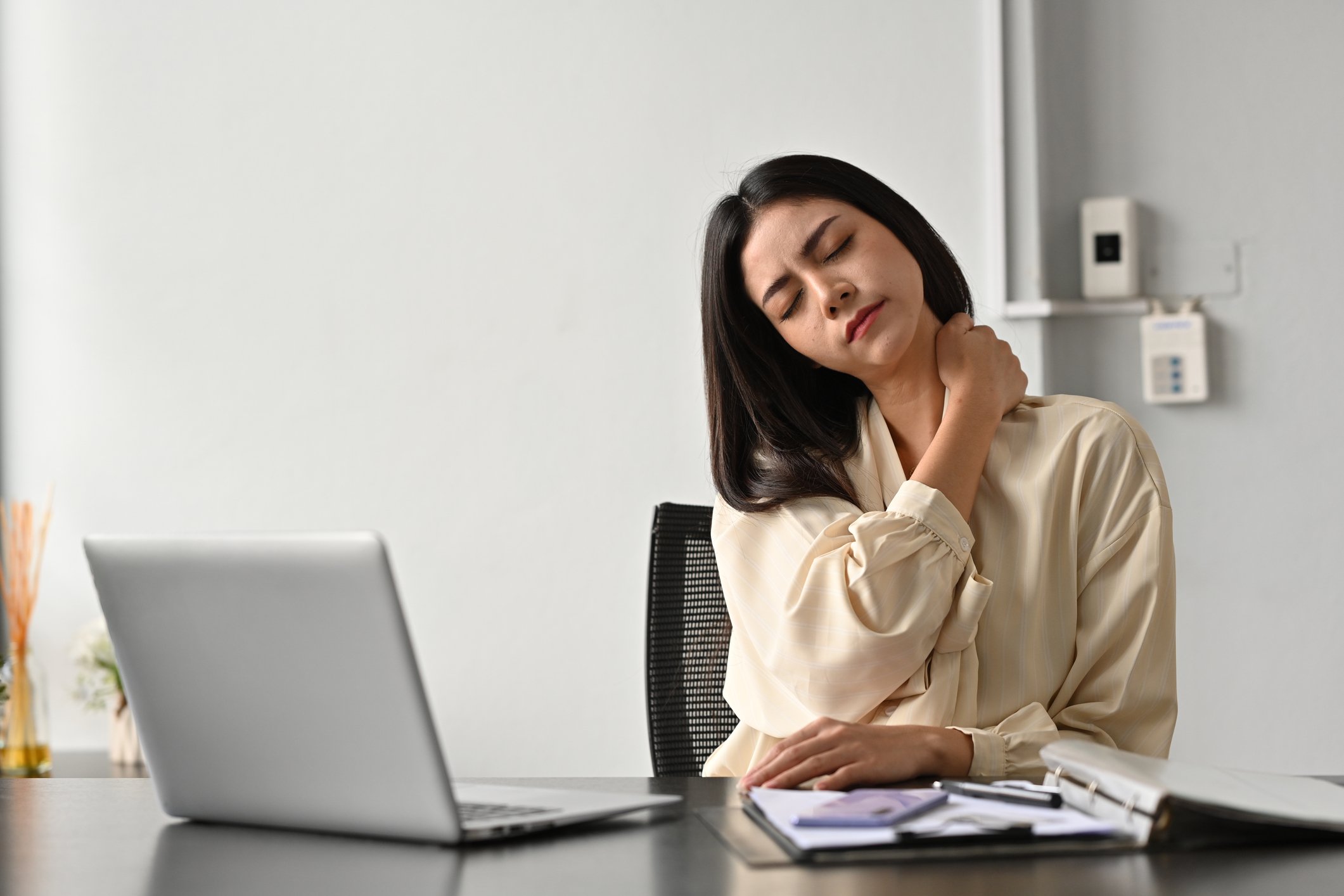 Woman sitting at a desk holding her neck in pain with a laptop and open notebooks.
