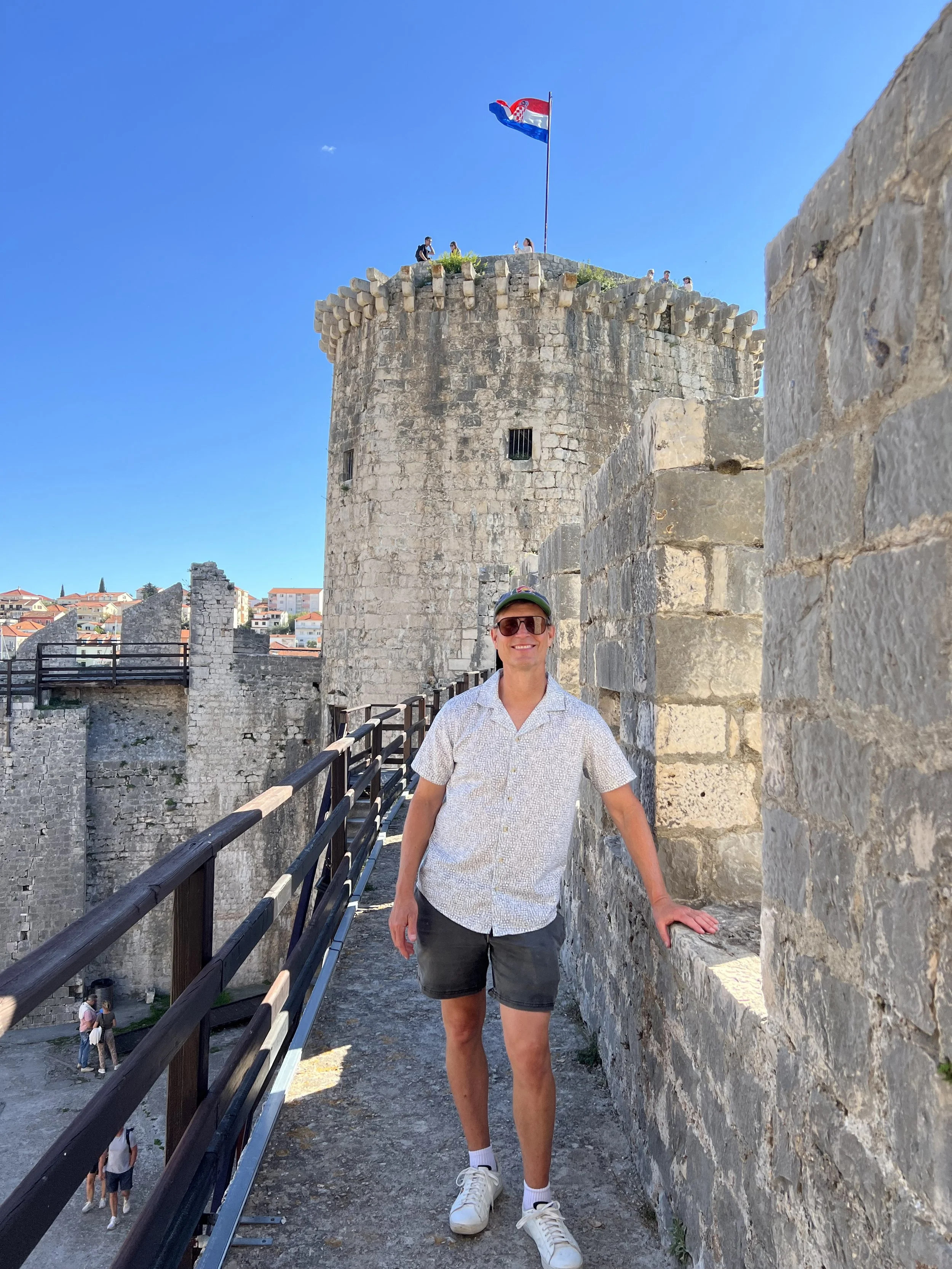 A man wearing sunglasses, a cap, a patterned short-sleeve shirt, and shorts, standing by a stone wall in front of a historic stone castle with a flag on top, under a bright blue sky.