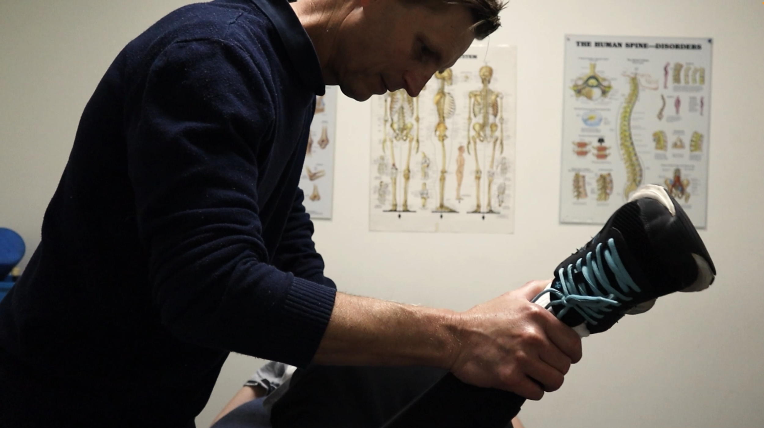 A man in a dark sweater applying a black and teal ice pack to a person's knee in a medical or physical therapy setting, with anatomical posters on the wall in the background.