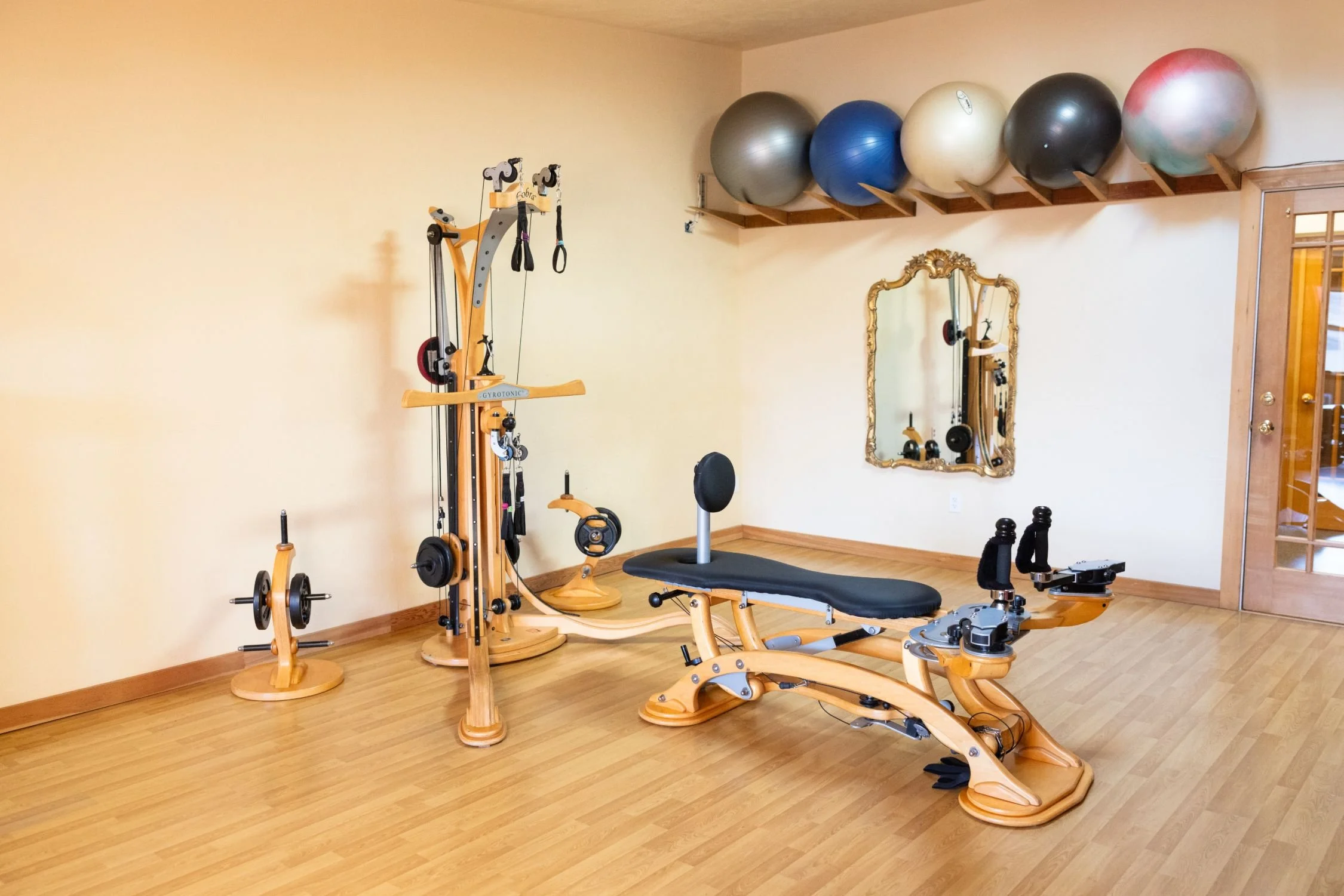 Home gym with wooden flooring, featuring a Pilates reformer machine, free weights, exercise balls on a wall-mounted shelf, and a decorative mirror
