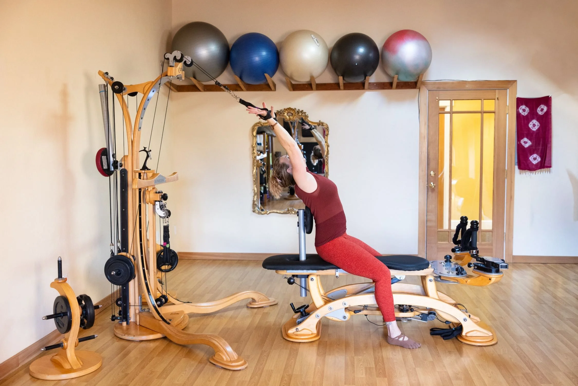 A woman doing a stretching exercise on a Pilates reformer machine in a home gym, with a mirror, exercise balls, and wooden furniture in the background.