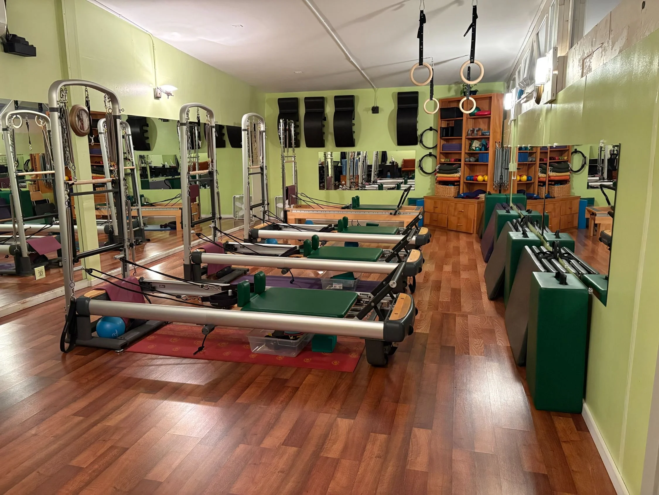 Pilates studio with reformer machines, mirrors, and exercise equipment, featuring light green walls and hardwood floors.