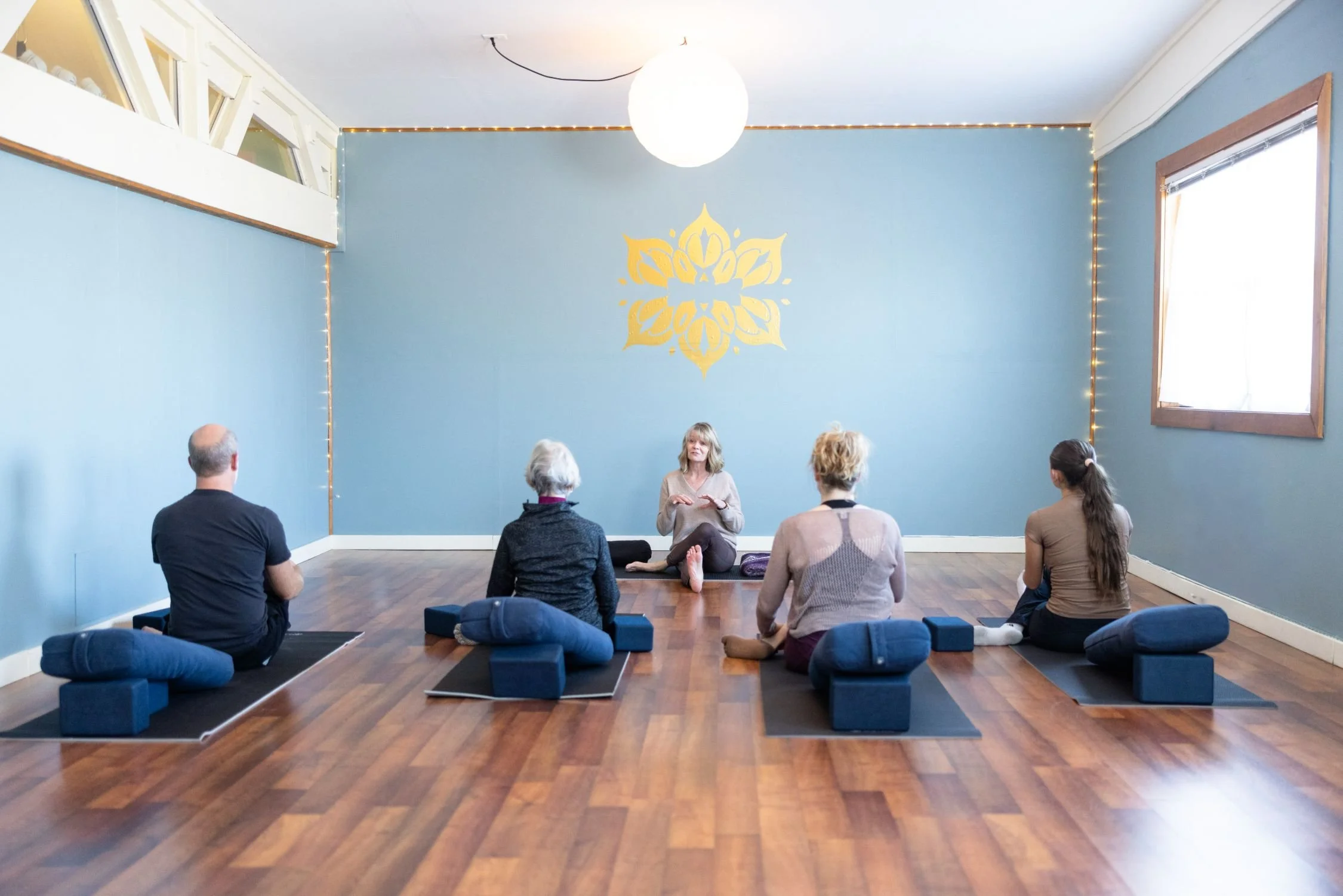 A yoga or meditation class with six participants seated on mats with bolsters in a bright studio, listening to an instructor sitting cross-legged at the front of the room.