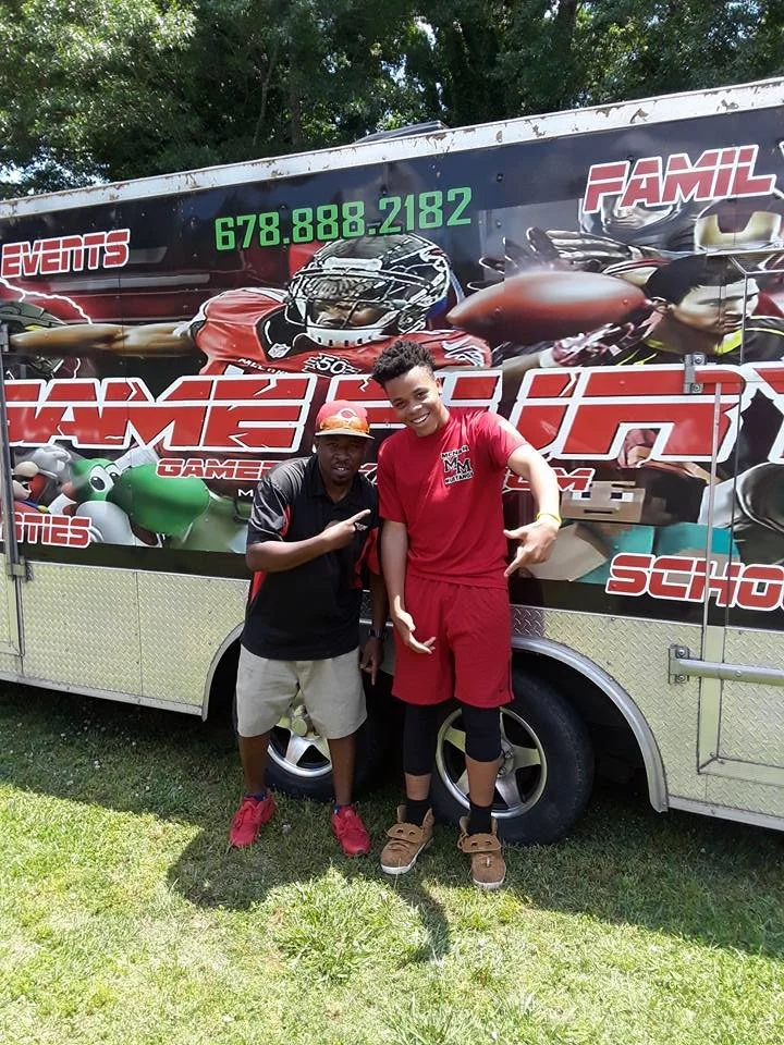 Two men posing in front of a colorful promotional trailer for a go-kart racing event. The trailer features images of racing cars and the words 'FAMIL', 'EVENTS', and a phone number. One man is wearing a black shirt and shorts, while the other is wearing a red shirt and shorts.