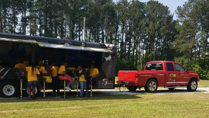 A group of people wearing yellow shirts gather around a food truck. There is a red pickup truck parked next to it on a grassy area with trees in the background.